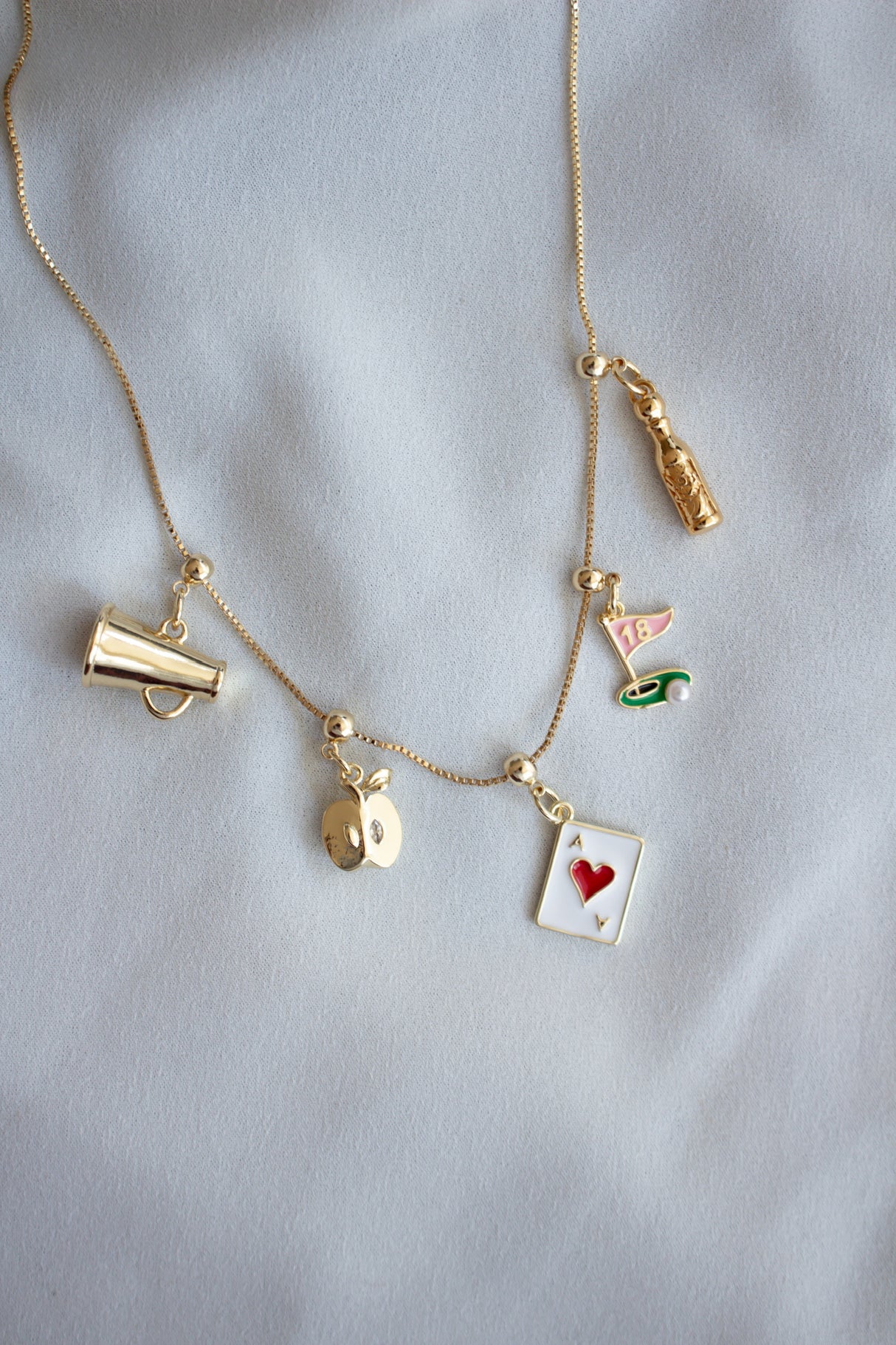 The Kinsey Designs Archives A+ Apple Grip Charm Bar Necklace, with fun charms like a megaphone, playing card, and bottle, makes a charming Teacher Appreciation Gift on light fabric.