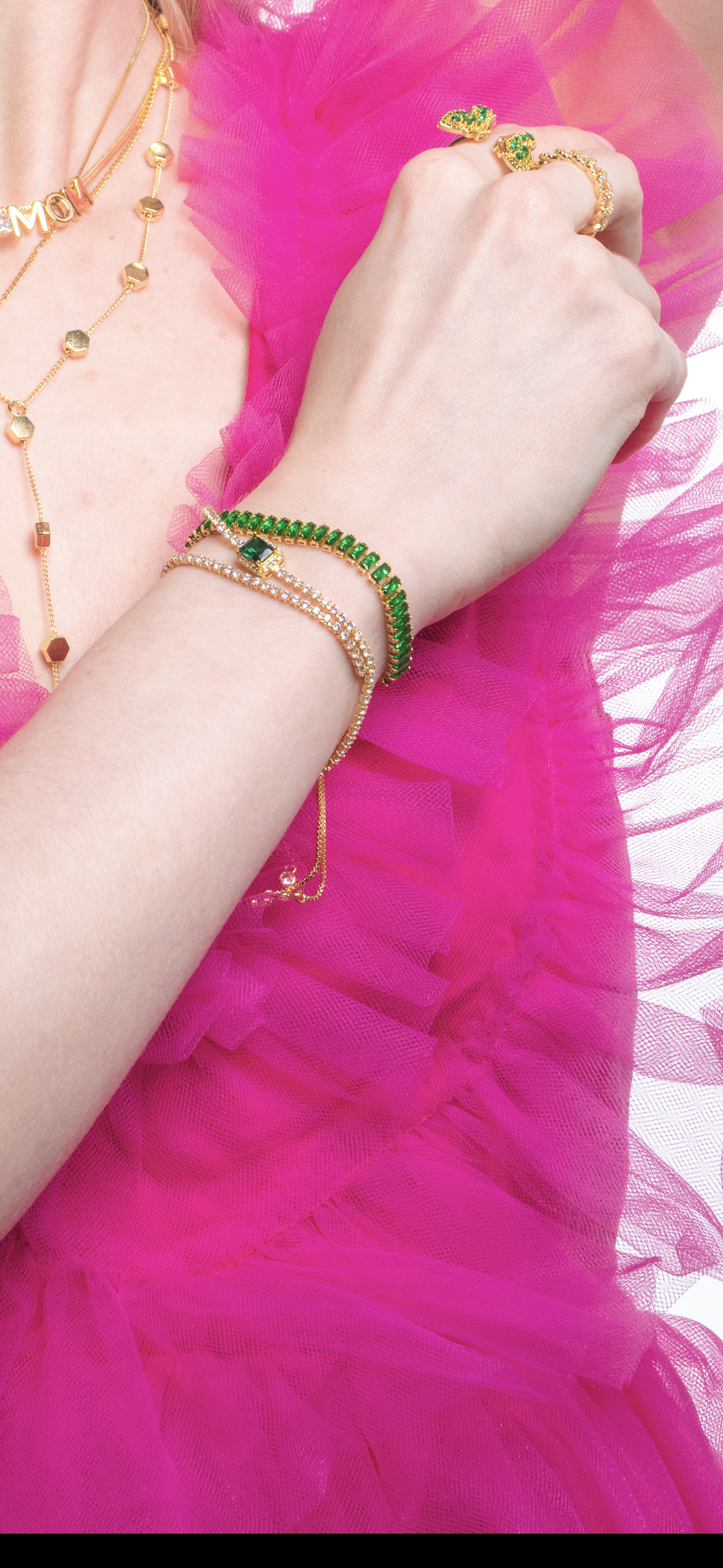A woman in a pink ruffled dress wears gold jewelry, including Kinsey Designs Archives’ Ivy Bracelet, along with green bracelets and gold rings set with green stones.