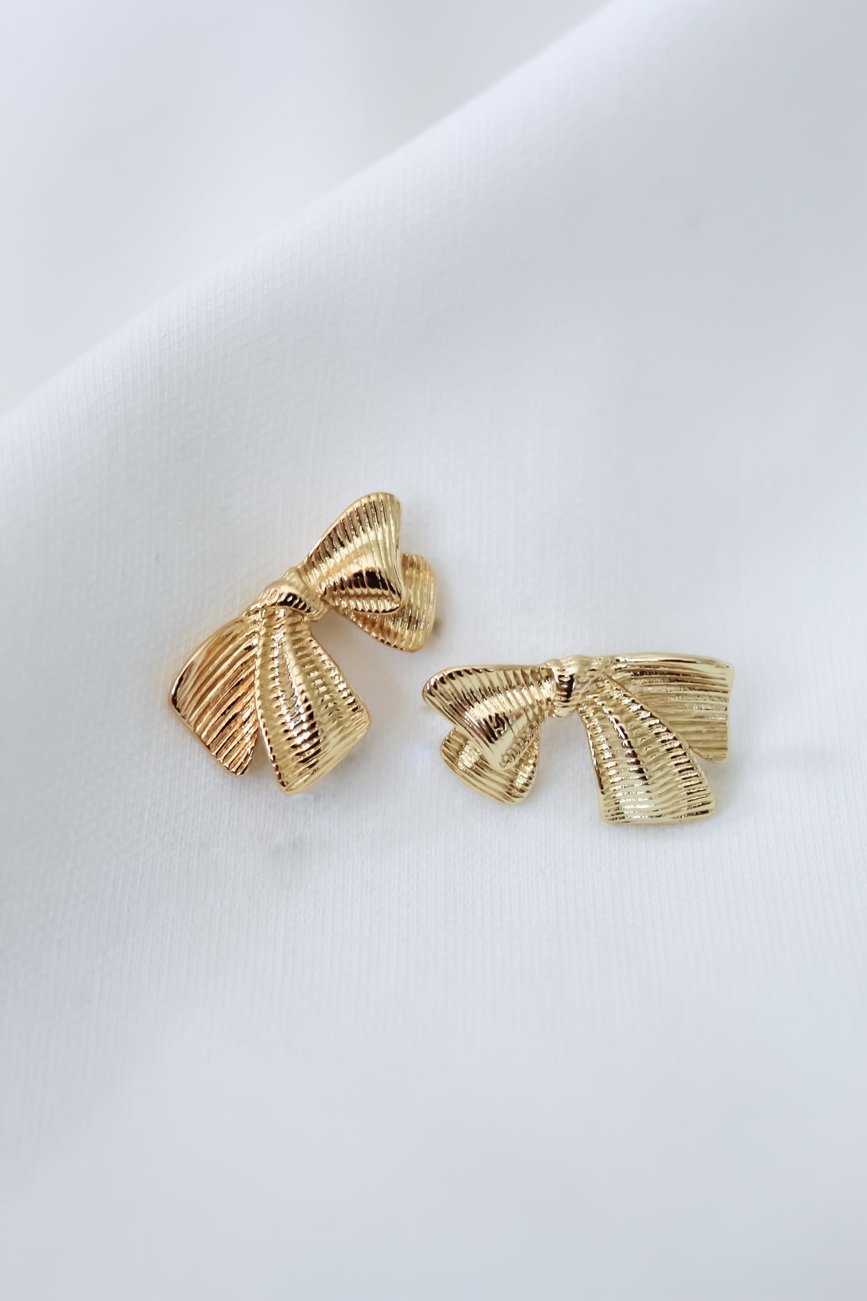 Enhance your look with the Izzy Bow Studs by Kinsey Designs—gold-tone bow earrings shown on a white fabric background.