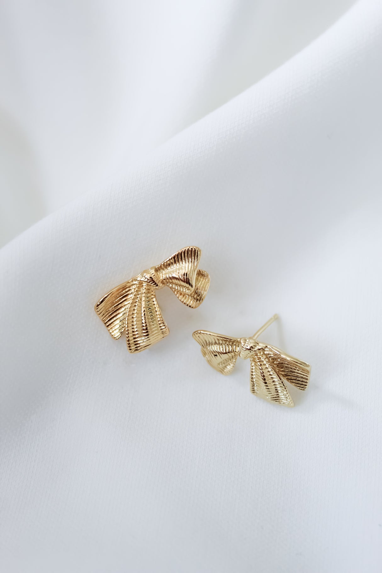 Gold-tone Izzy Bow Studs by Kinsey Designs, displayed on white fabric, add a touch of elegance to any outfit.