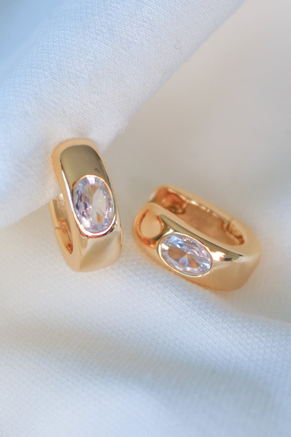 The Jack Huggie by Kinsey Designs features gold-filled earrings with oval clear gemstones, displayed on soft white fabric. These sample sale pieces add an elegant touch to any look.