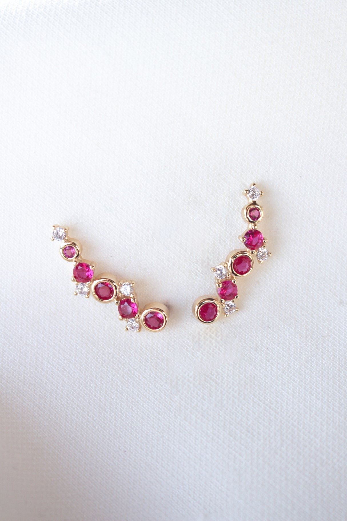 The Jax Climber Fuchsia earrings by Kinsey Designs feature gold filled curves adorned with vibrant fuchsia gemstones and sparkling cubic zirconia, beautifully displayed on a white fabric background.