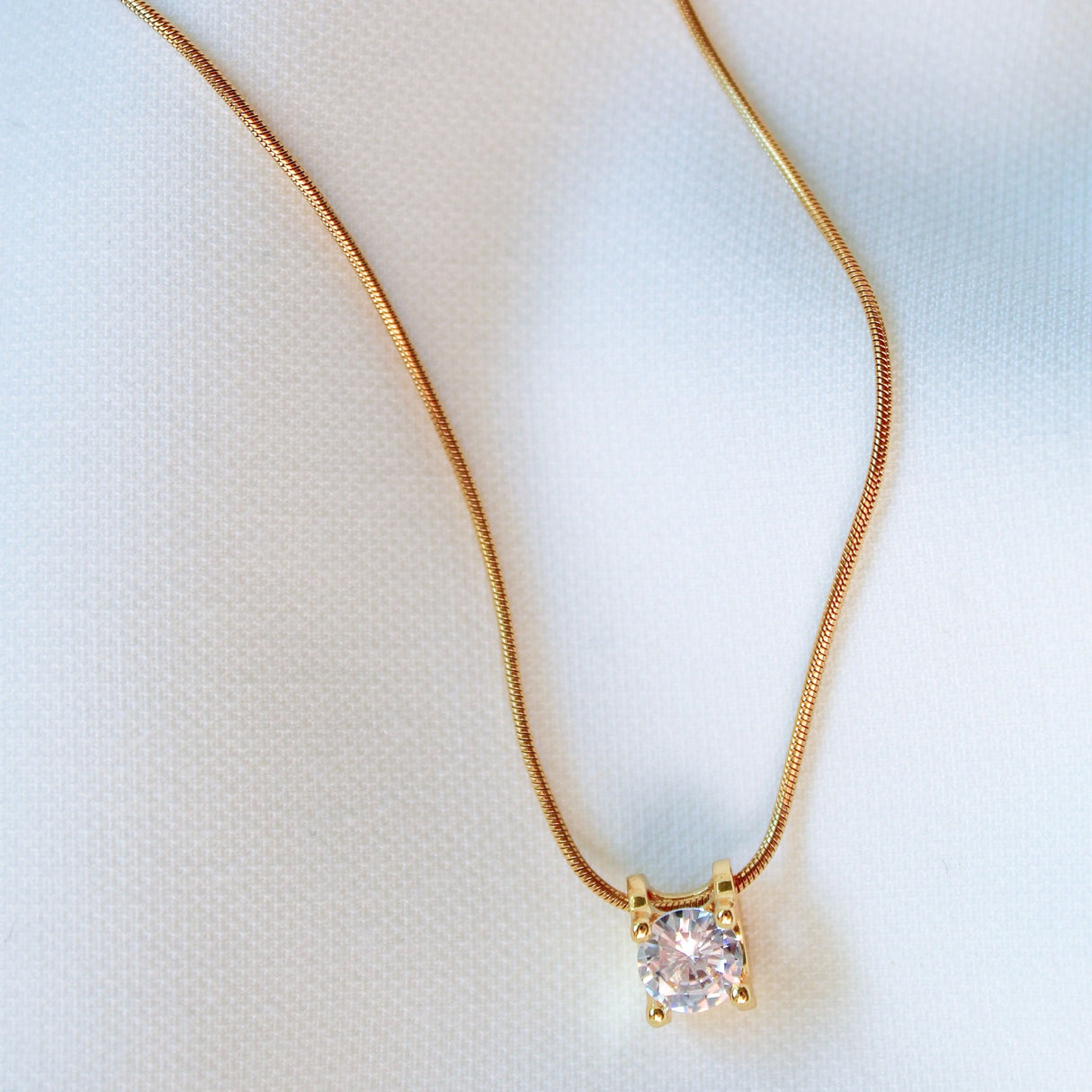 The Jenna Necklace by Kinsey Designs Archives features an 18-inch gold filled chain with a square pendant set with a single sparkling gemstone, displayed on a white fabric background for an elegant touch.