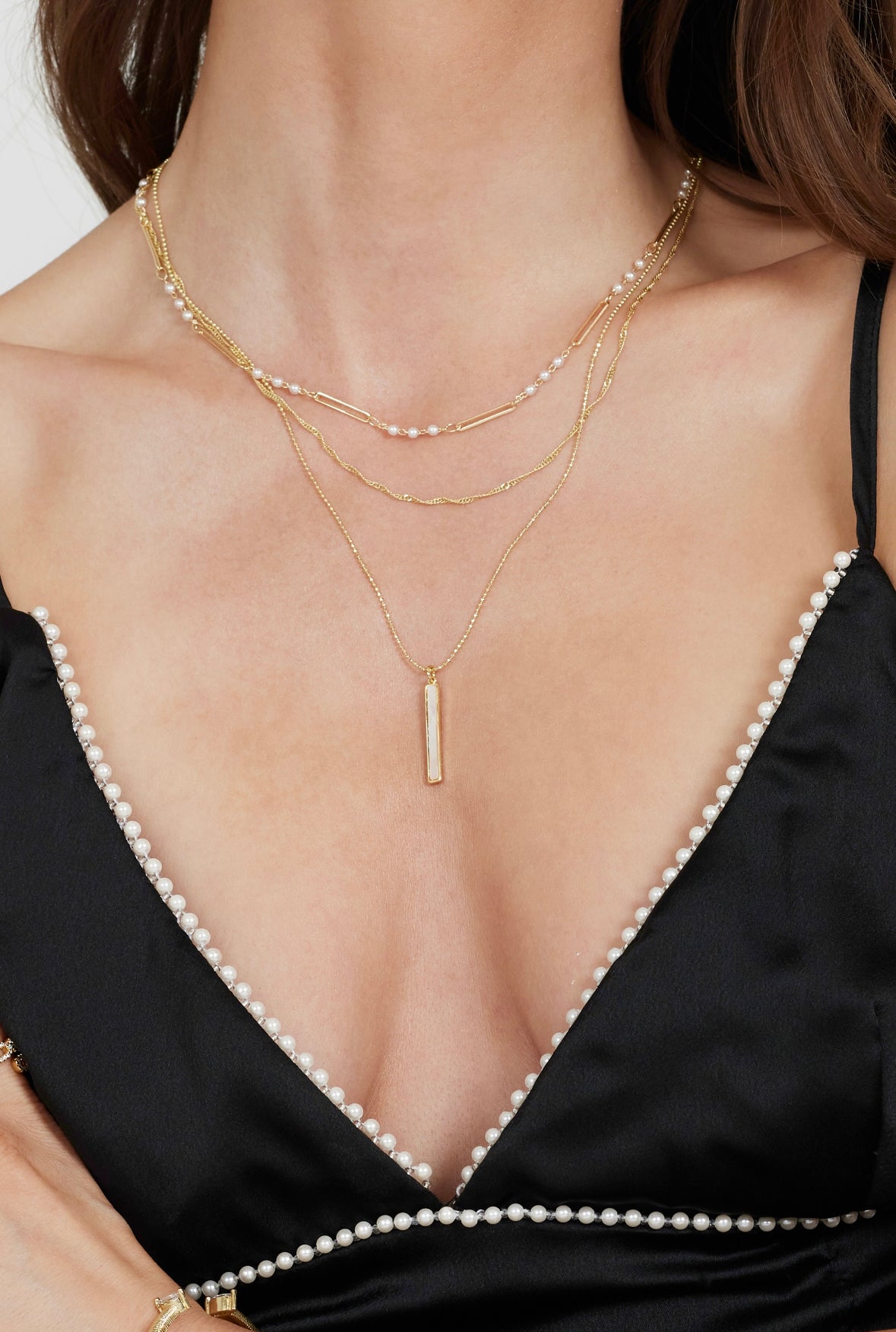 A woman wears the Kinsey Designs Archives Kalani Pearl Necklace and layered chains with a black dress featuring mother-of-pearl trim along the neckline.