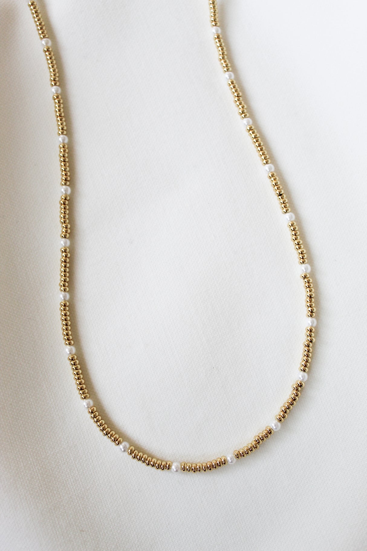 The Kandi Necklace by Kinsey Designs, a gold and white beaded 16" piece with clasp and extender, is displayed on a white fabric background.