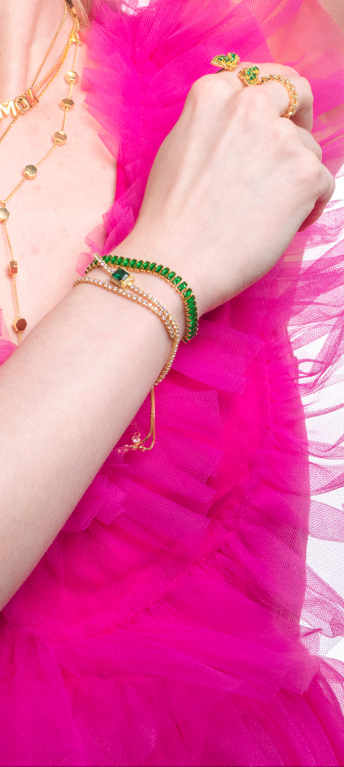 A woman in a pink ruffled dress wears gold jewelry, green gemstone rings, and the Kinsey Designs Katie Bracelet—a gold filled bracelet adorned with cubic zirconia.