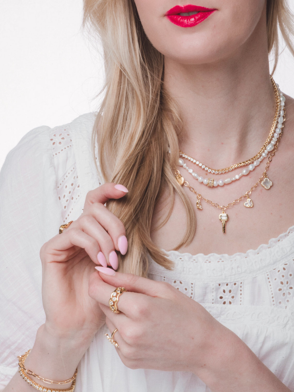 Woman in white dress wears layered Kinsey Designs gold-filled necklaces, featuring the Key Hearts Charm Necklace, with pink nails and bright pink lipstick, touching her hair.