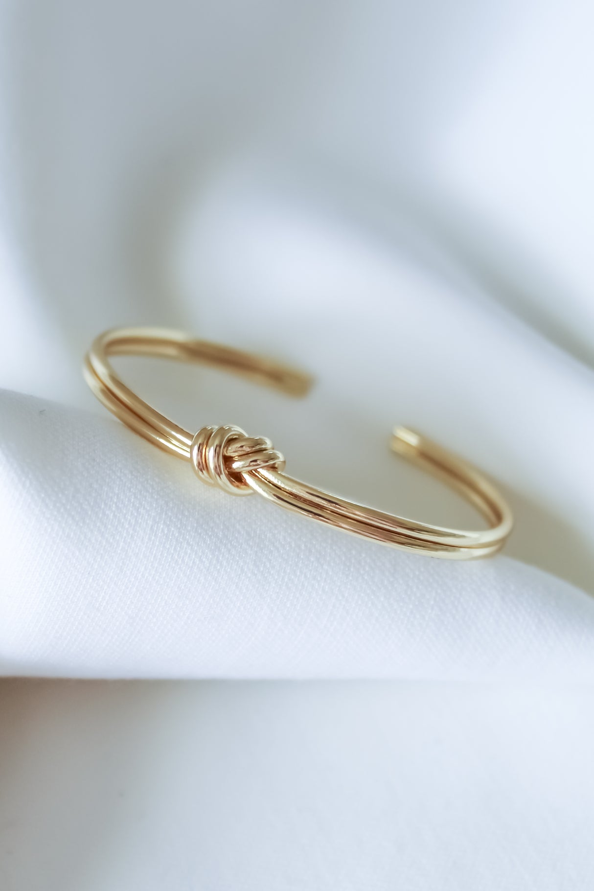 The Knotty Cuff Bracelet by Kinsey Designs features a delicate knot motif, elegantly displayed on white fabric—an ideal bridal gift or a chic accessory for any occasion.