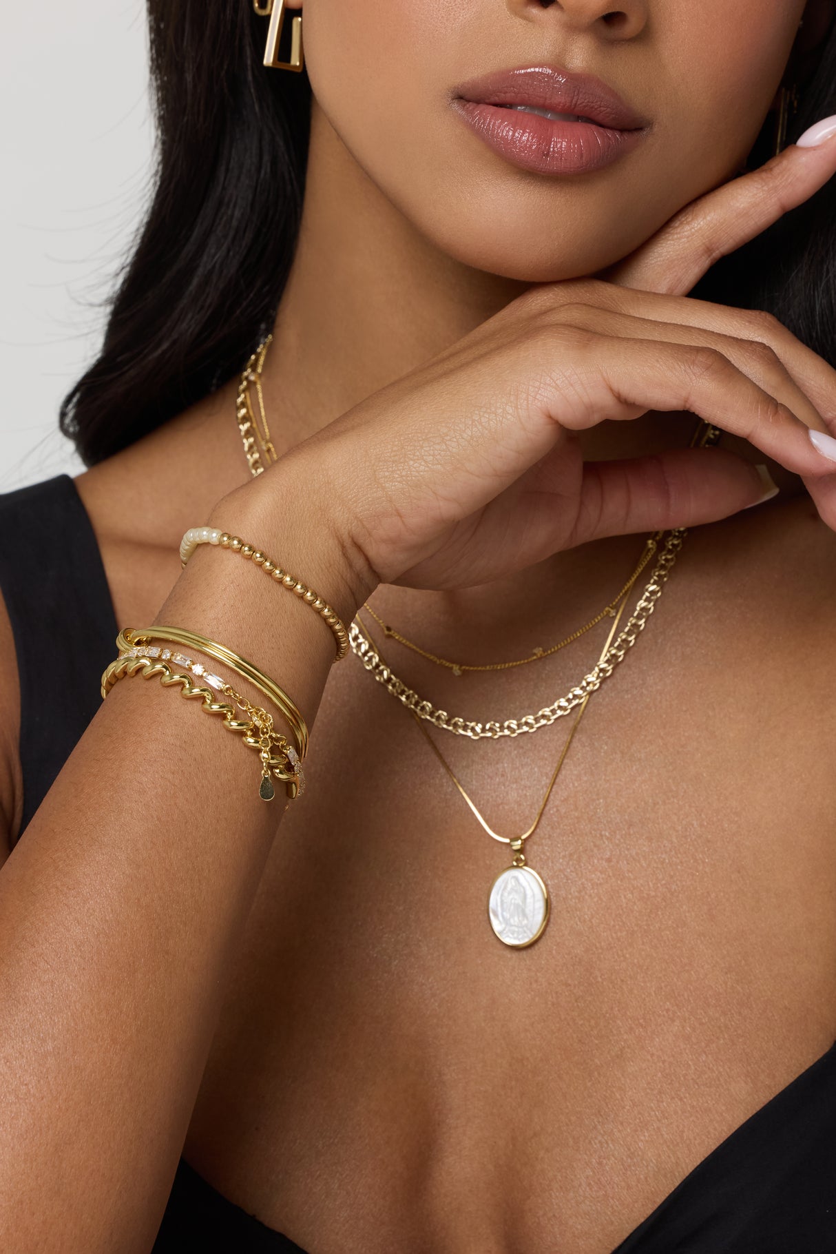A woman wears layered gold necklaces, earrings, and the Kinsey Designs Knotty Cuff Bracelet with a knot motif—an elegant bridal gift—as her hand rests gracefully near her chin.