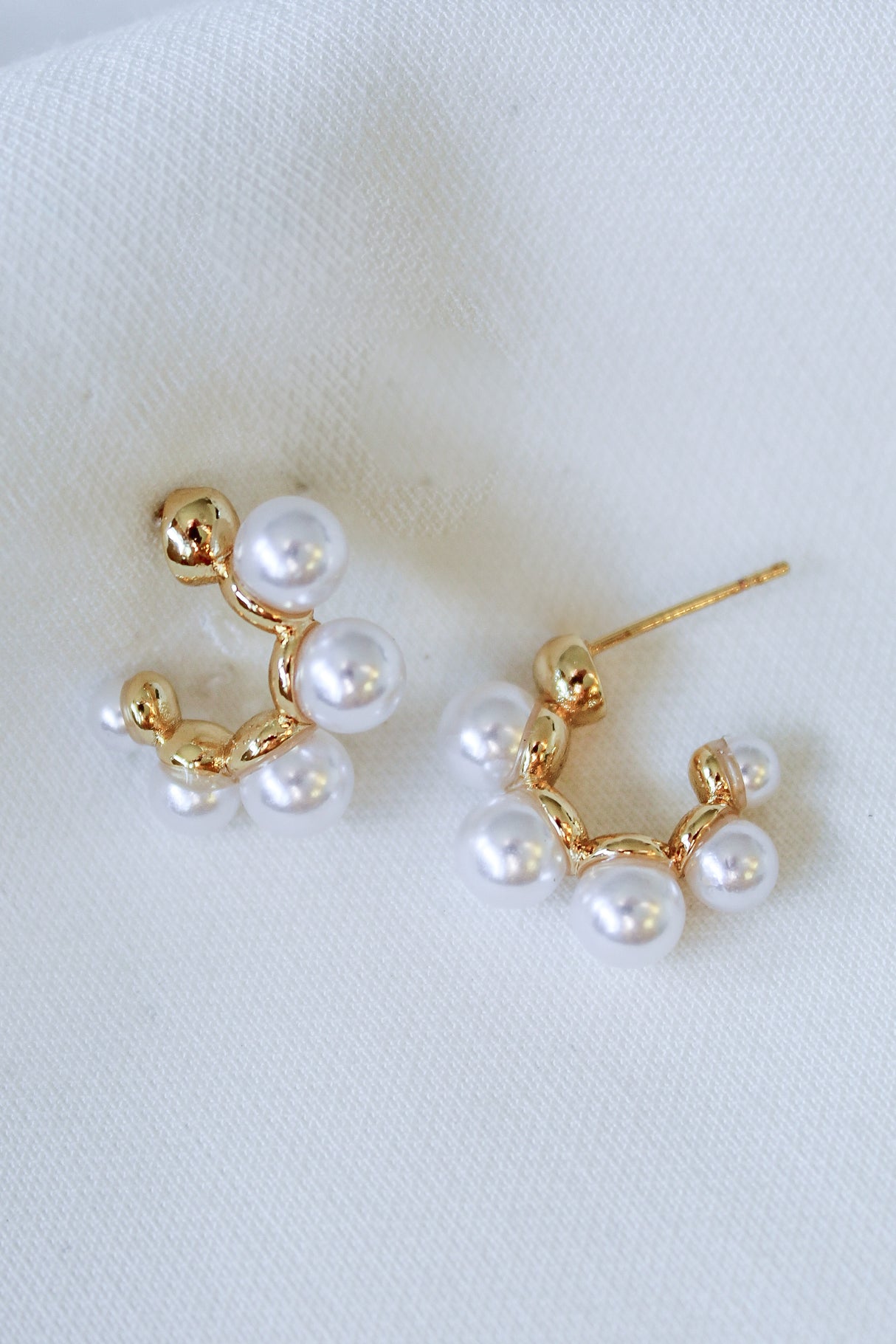 The Kona Pearl Hoops by Kinsey Designs are gold earrings adorned with clusters of freshwater pearls, shown on a white textured fabric background.