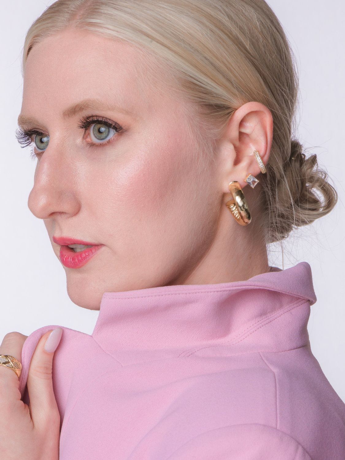 A woman with blonde hair in a low bun, wearing Kinsey Designs' Ladrielle Hoop gold-filled earrings and a pink high-collar jacket, looks to the left.
