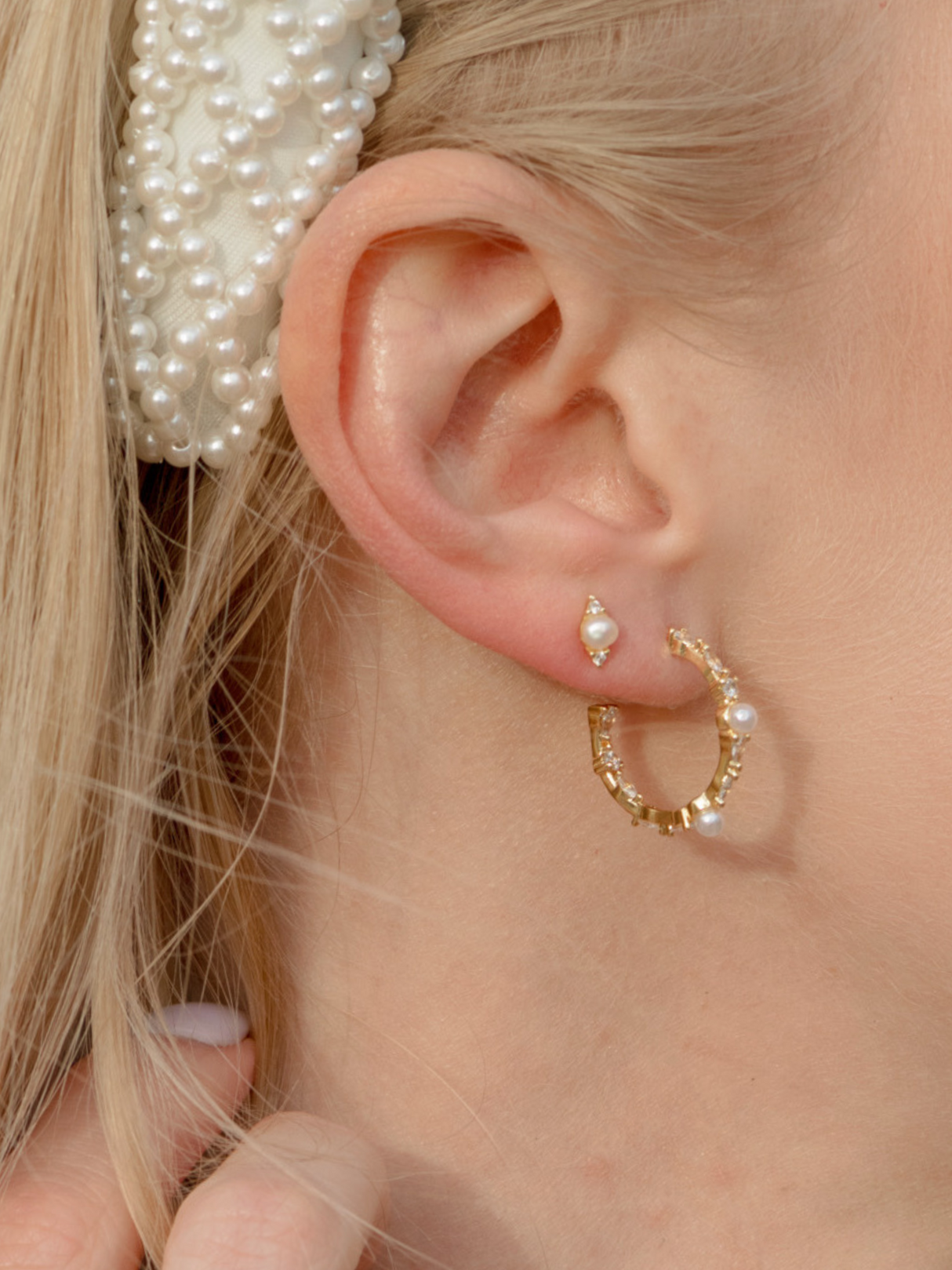 Close-up of a woman's ear adorned with the Lollita Pearl Hoop by Kinsey Designs, paired with a pearl headband and pearl stud for a chic, layered look.