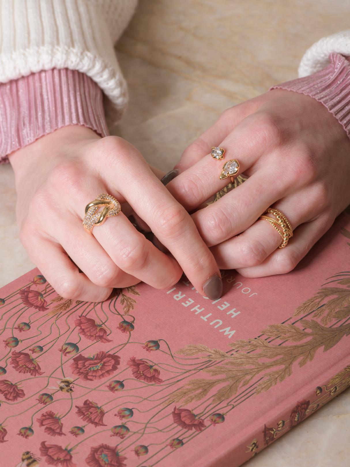 Hands adorned with the Lorelai Wrap Ring by Kinsey Designs and gold bands rest on a pink floral book; person wears a white and pink sweater.