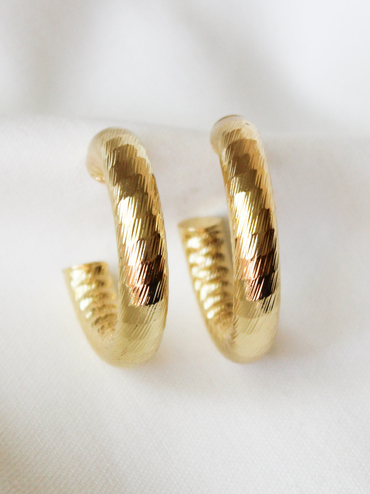 The Kinsey Designs Ladrielle Hoop, a pair of textured gold filled earrings, rests on a white fabric background.