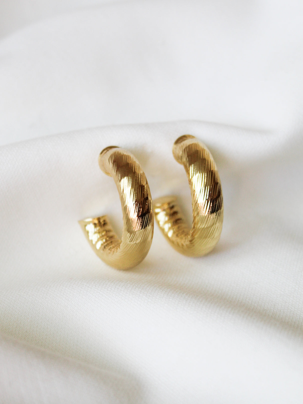 The Ladrielle Hoop by Kinsey Designs, a pair of textured gold filled earrings, rests on white fabric. Their classic small hoop design brings an elegant touch to any look.