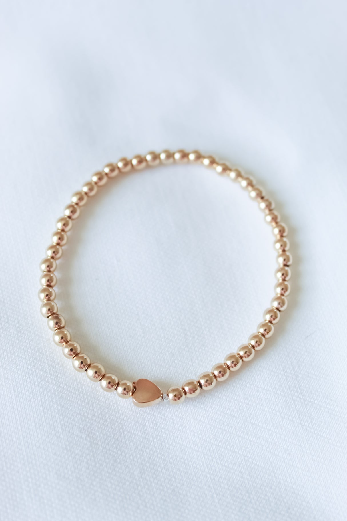 The Liam Heart Bracelet by Kinsey Designs features gold-filled beads and a small heart charm on a white fabric background. With its adjustable stretch design, this bracelet combines elegance and comfort.