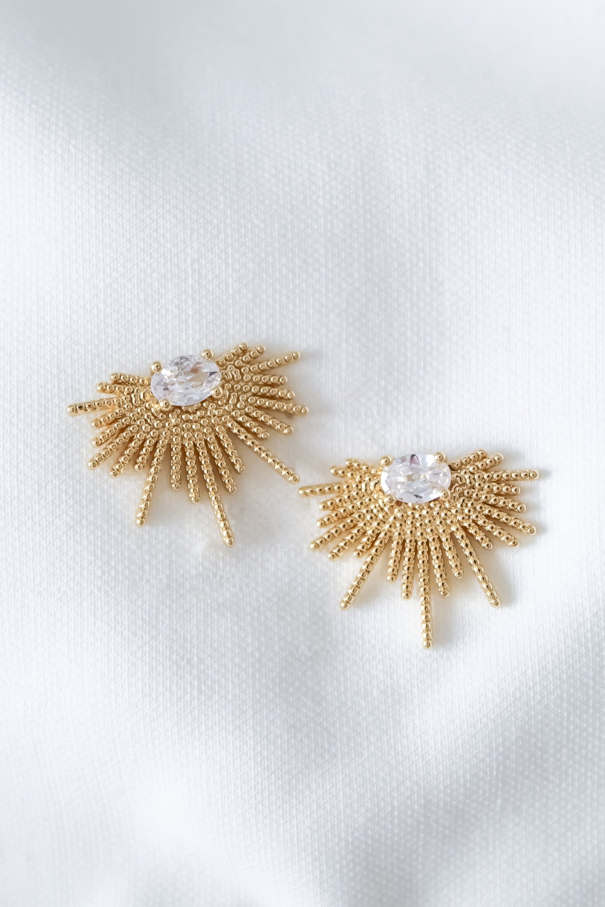 The Linga Earring by Kinsey Designs features gold filled sunburst accents with a central clear gemstone, shown on a white textured fabric background. Final sale.