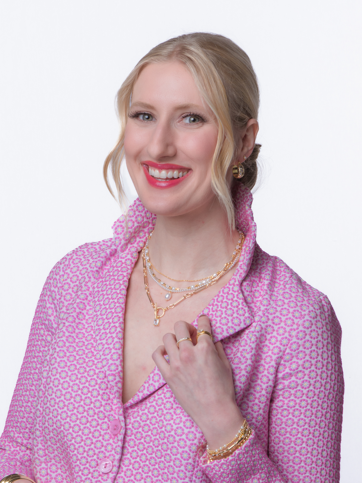 Smiling woman in a pink patterned jacket wears layered gold jewelry, including the elegant Louie Pearl Necklace by Kinsey Designs, against a white background.