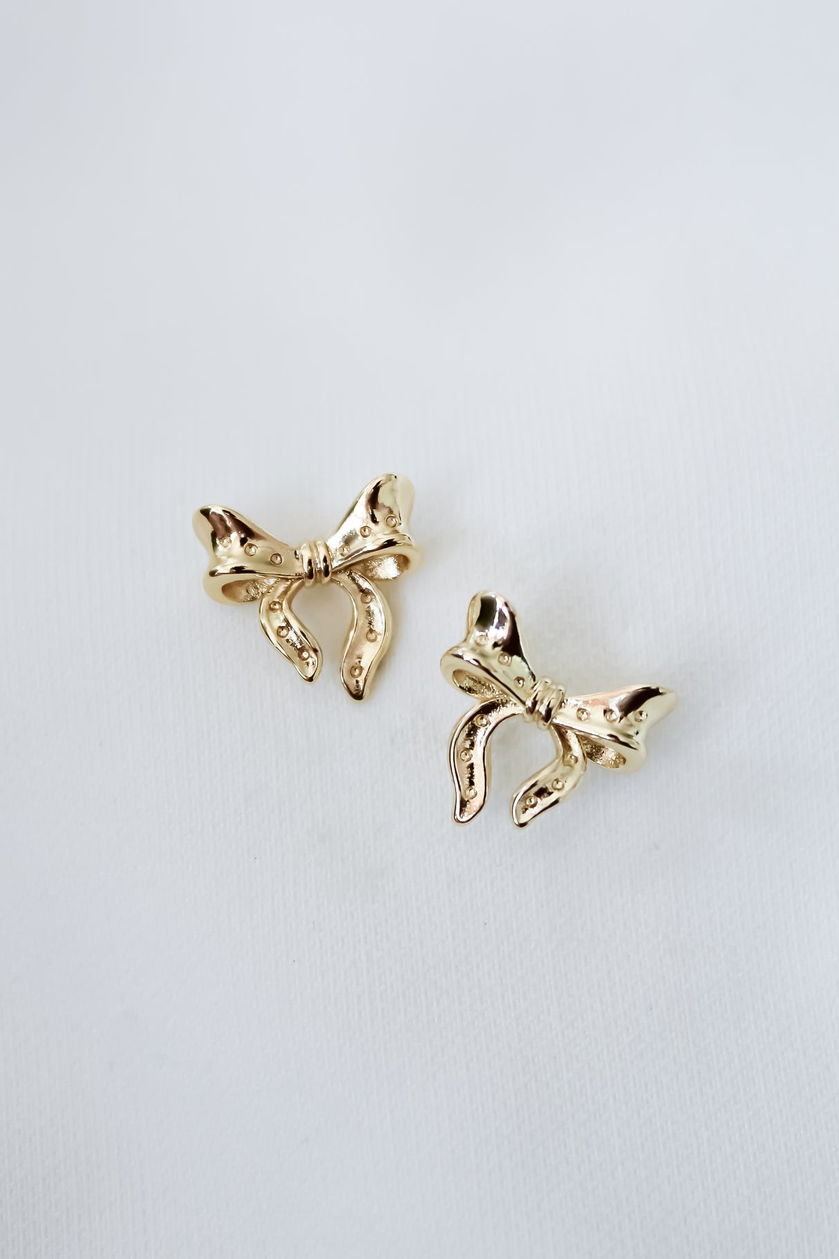 The Lova Studs by Kinsey Designs, featuring two shiny gold bows, make perfect feminine jewelry and are elegantly displayed against a plain white background.