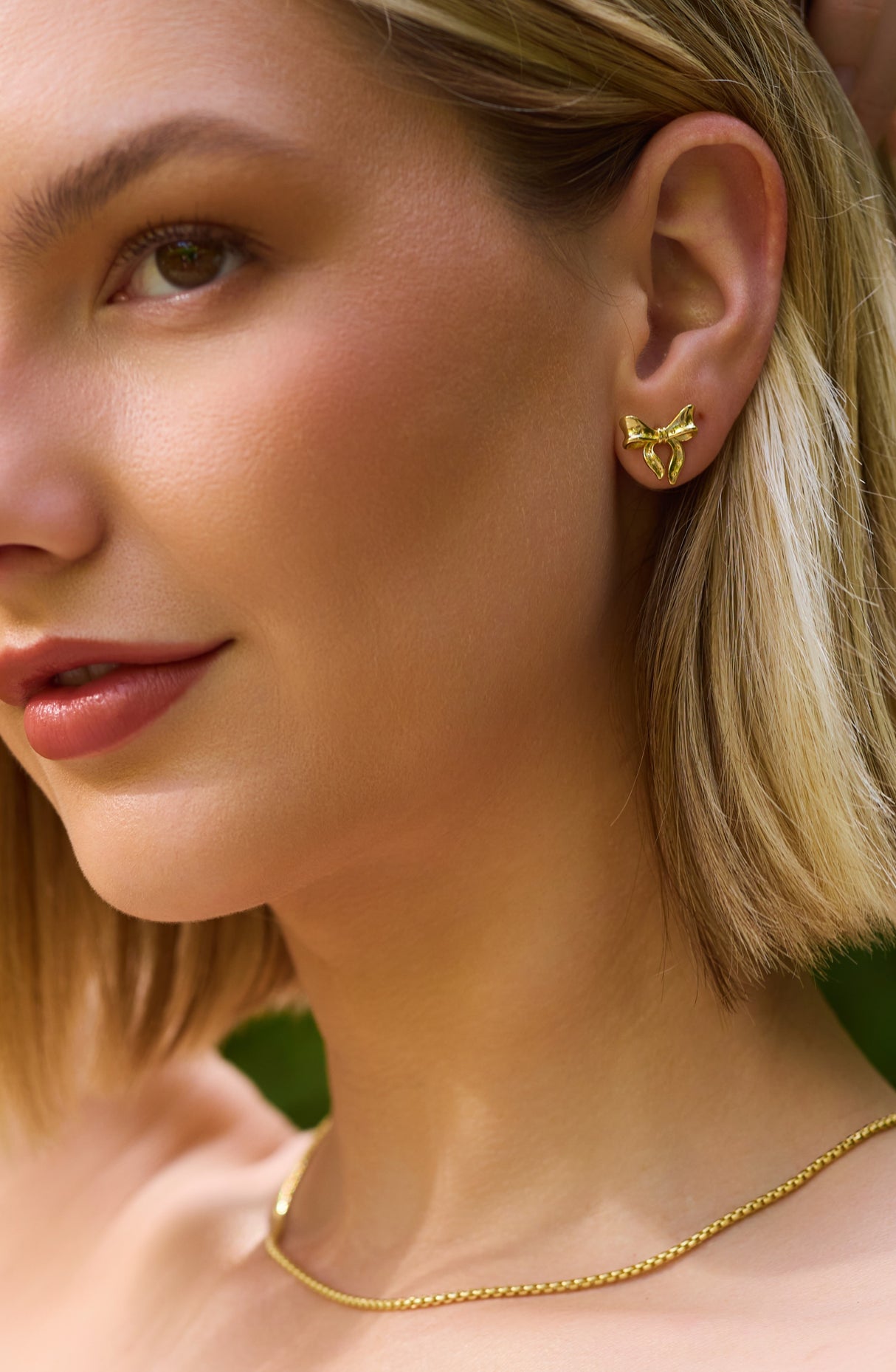 A woman with short blonde hair wears Kinsey Designs' Lova Studs gold bow earrings and a gold chain necklace.