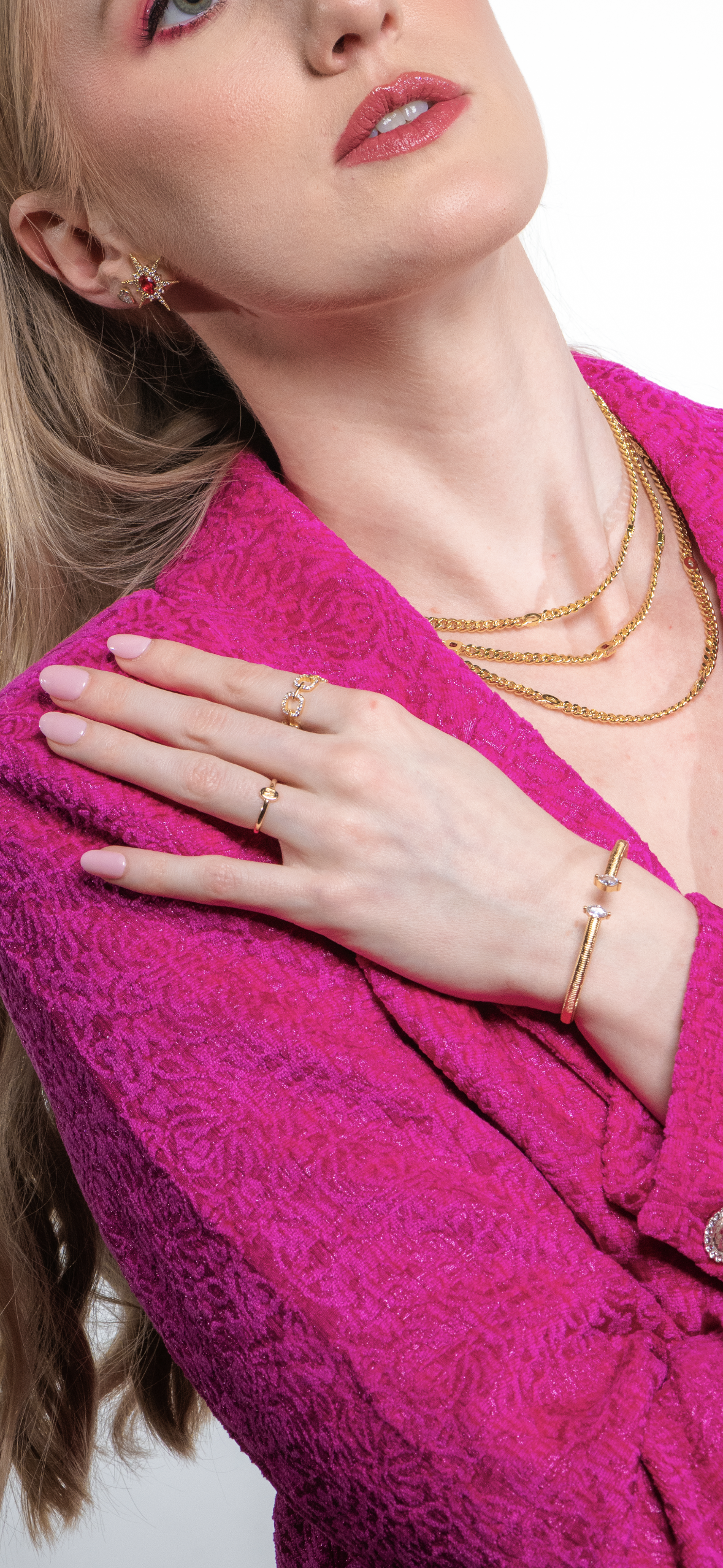 A woman in a textured pink blazer, hand on collar, highlights the Kinsey Designs Lover Necklace—a delicate gold-filled piece—layered with other chains and paired with light pink nail polish.