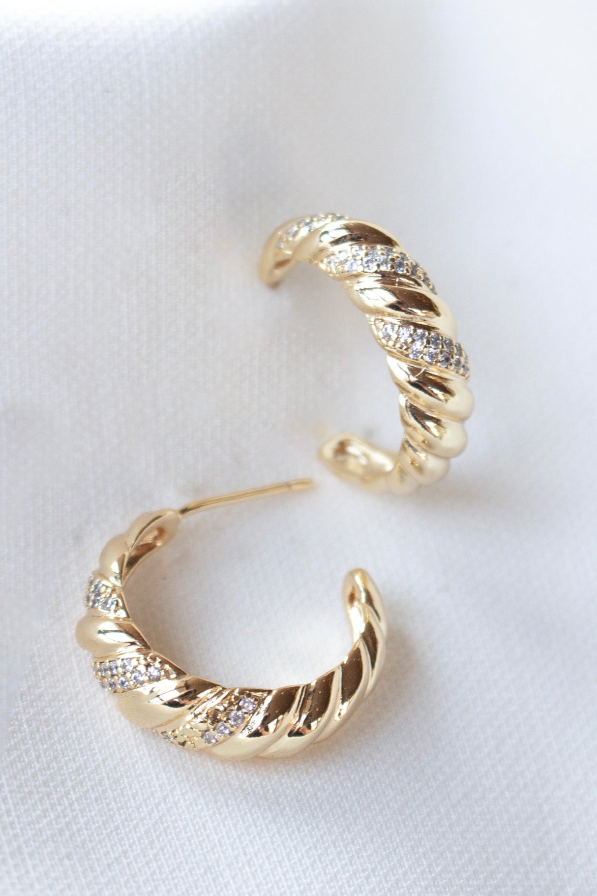 The Luca Hoop by Kinsey Designs Archives features gold filled twisted hoops with small cubic zirconia crystals, shown on a white fabric background. This stylish earring adds shimmer to your look.