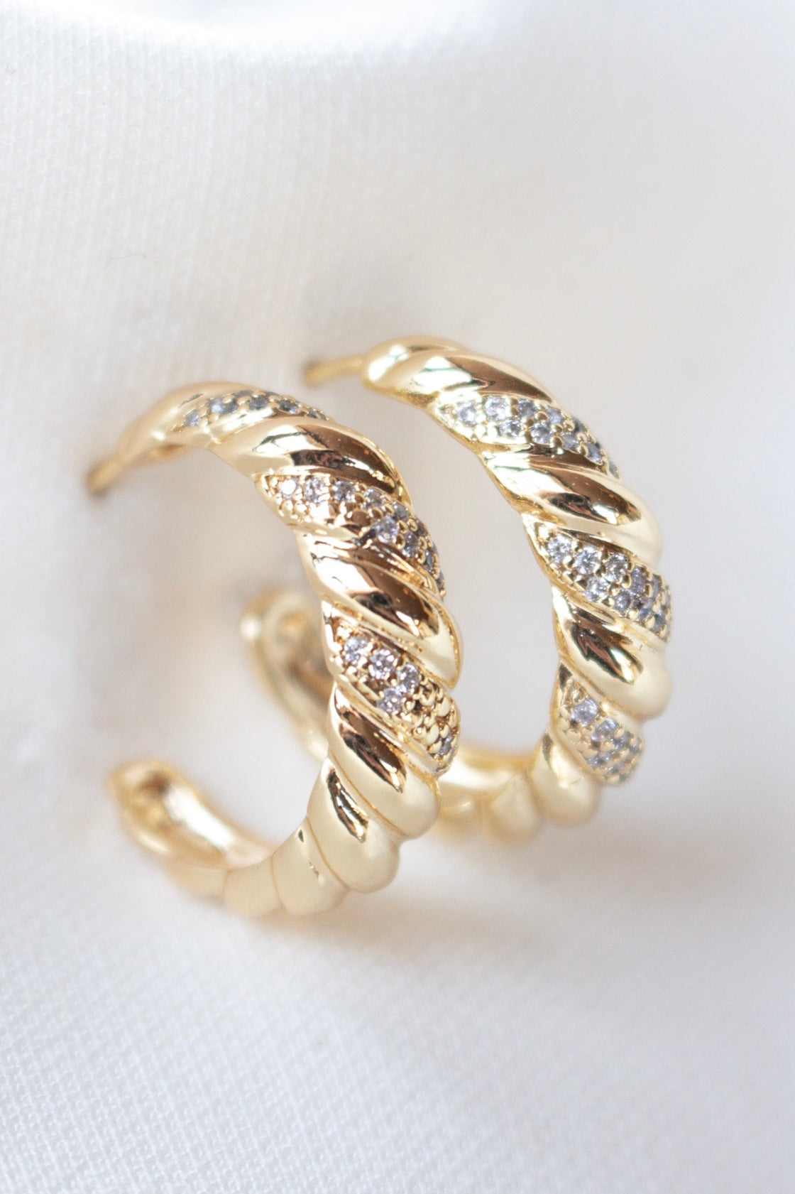 The Luca Hoop by Kinsey Designs Archives features gold-filled twisted hoops with alternating smooth and pavé cubic zirconia sections, shown on a white fabric background.