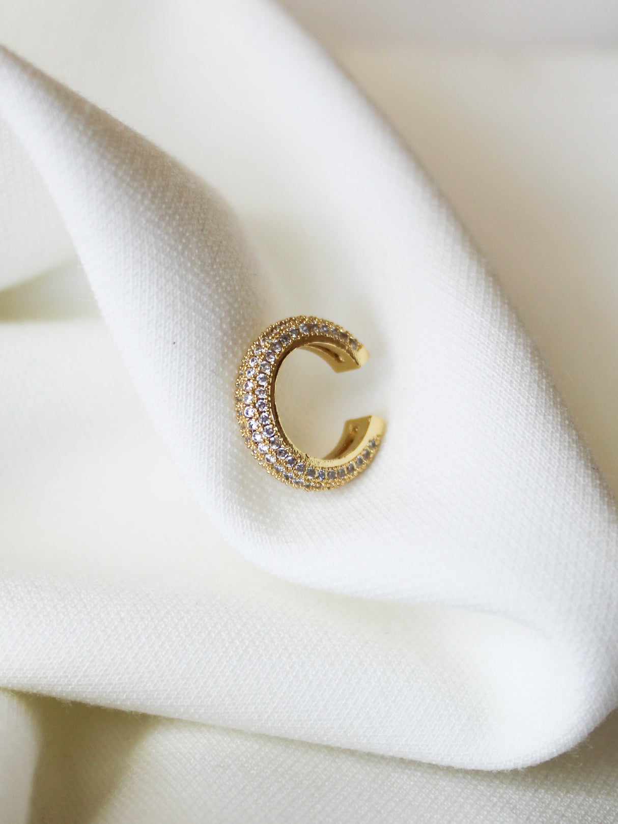 The Luce Ear Cuff by Kinsey Designs is a gold-filled, crescent-shaped earring adorned with small crystals and a 0.5" circumference, elegantly displayed on folded white fabric.