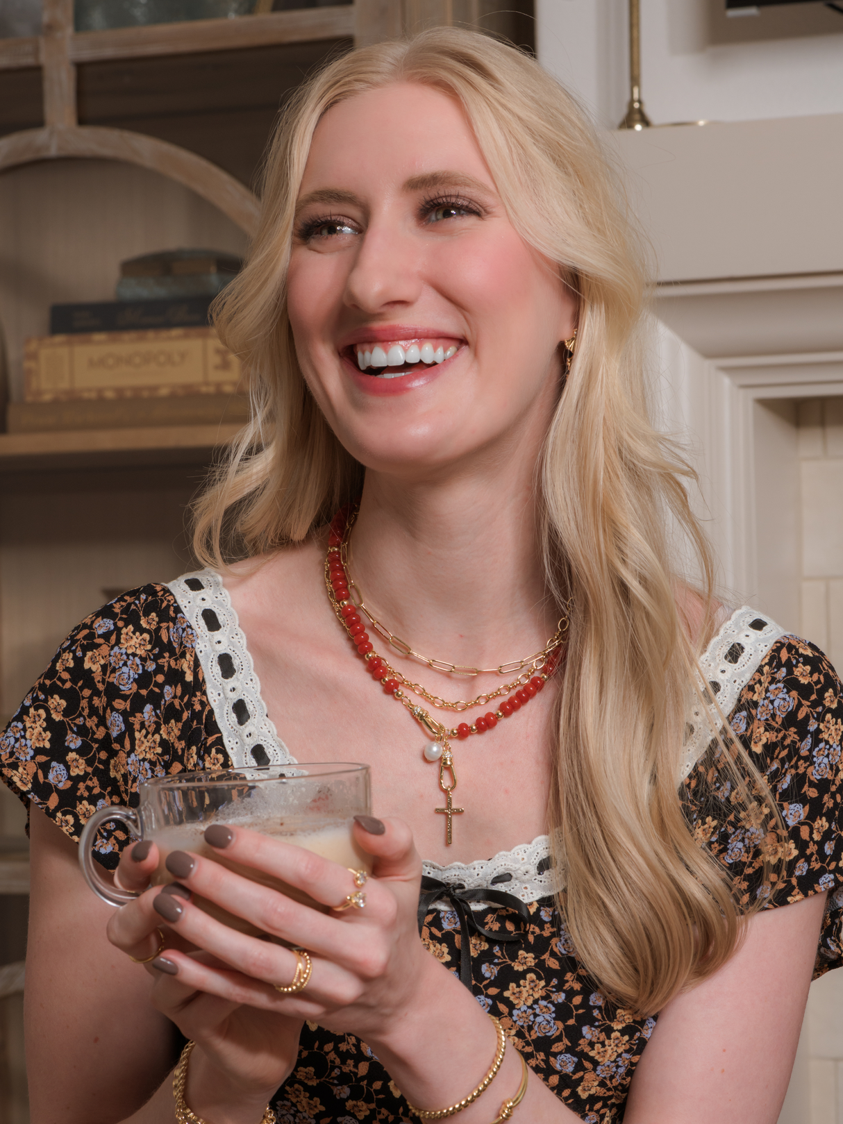 Smiling woman with long blonde hair, in a floral dress and layered gold filled necklaces, including the delicate Kinsey Designs Maeve Necklace and a paperclip necklace, holding a glass cup.