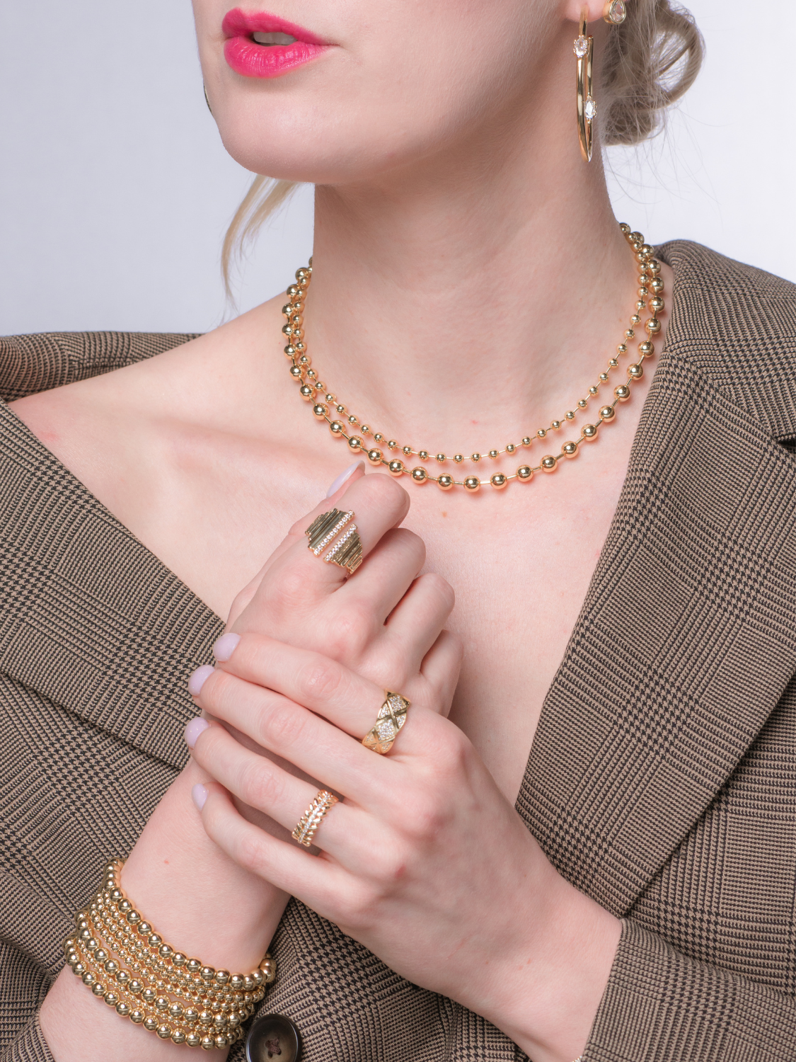 Woman in a plaid blazer wearing gold jewelry, including a necklace, bracelet, hoop earrings, and the Mallon Ring by Kinsey Designs with a quilted grid pattern.