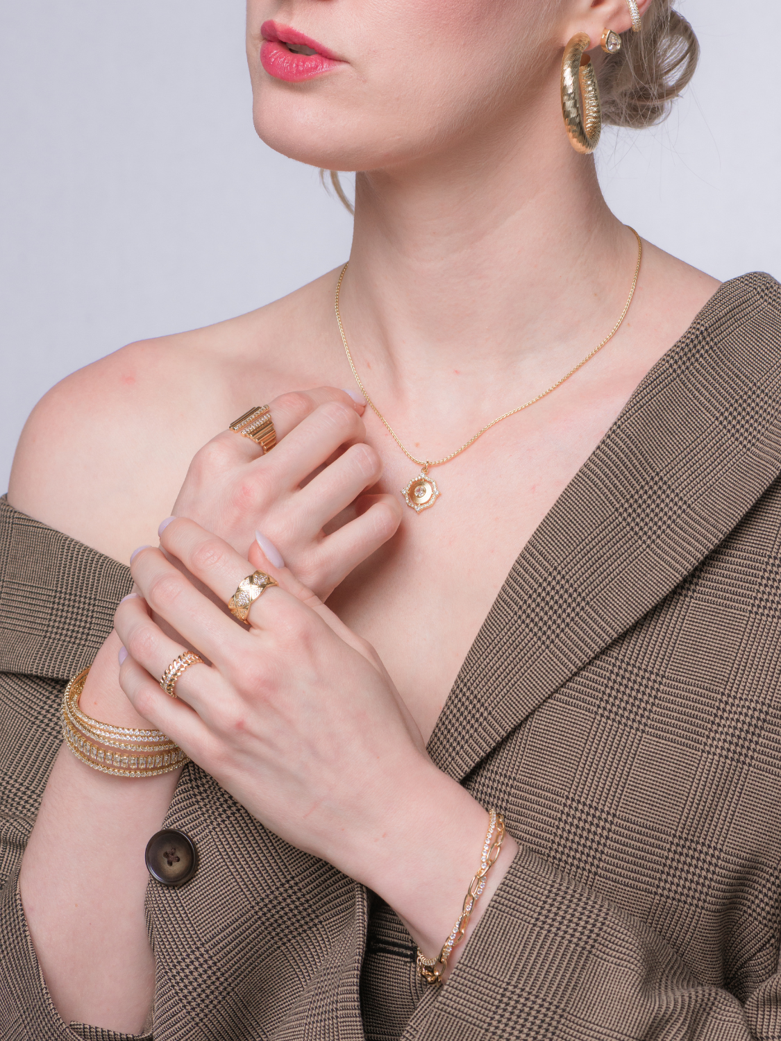 A woman in a plaid blazer models gold jewelry, featuring rings, a necklace, earrings, and the stunning Marc Tennis Bracelet by Kinsey Designs.