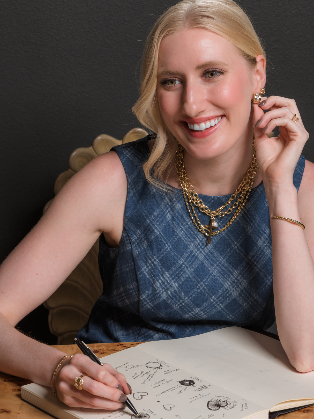 A smiling woman in a blue dress sketches jewelry designs in her notebook, wearing the Kinsey Designs Marigold Cross Wrap Necklace.