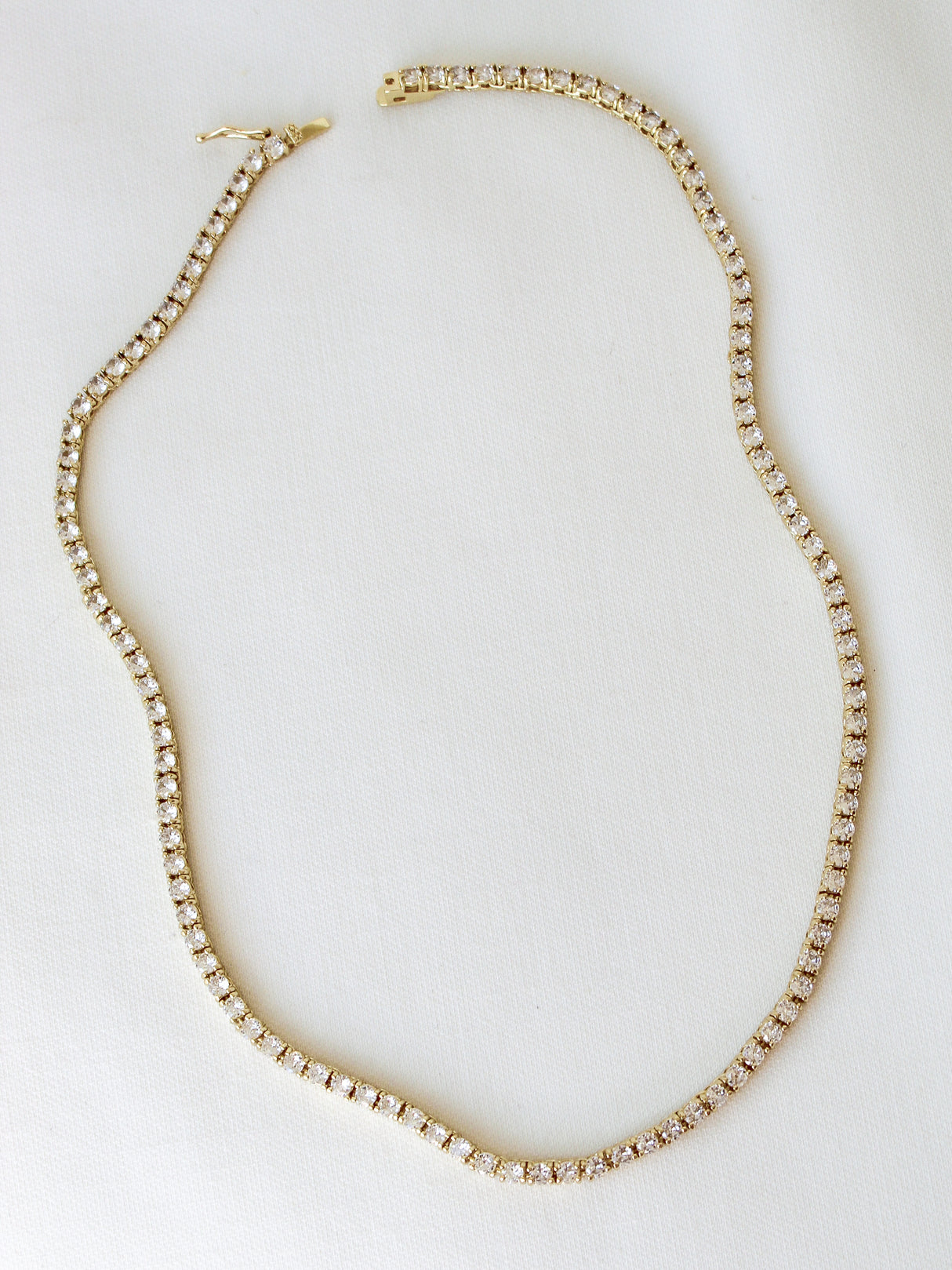 The Malia Tennis Necklace by Kinsey Designs Archives features small, round white stones set in gold fill and is displayed on a white fabric. This elegant 15" necklace suits any occasion.