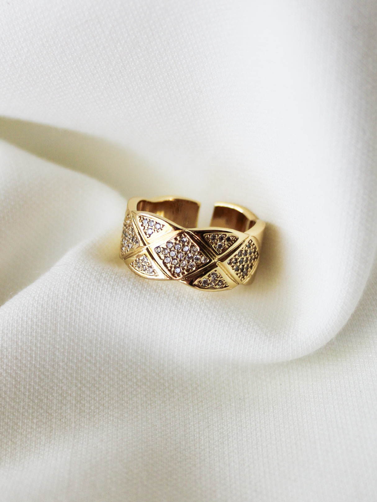 The Mallon Ring by Kinsey Designs features a gold band with a crisscross quilted grid pattern and small pavé stones, showcased on a soft white fabric background.