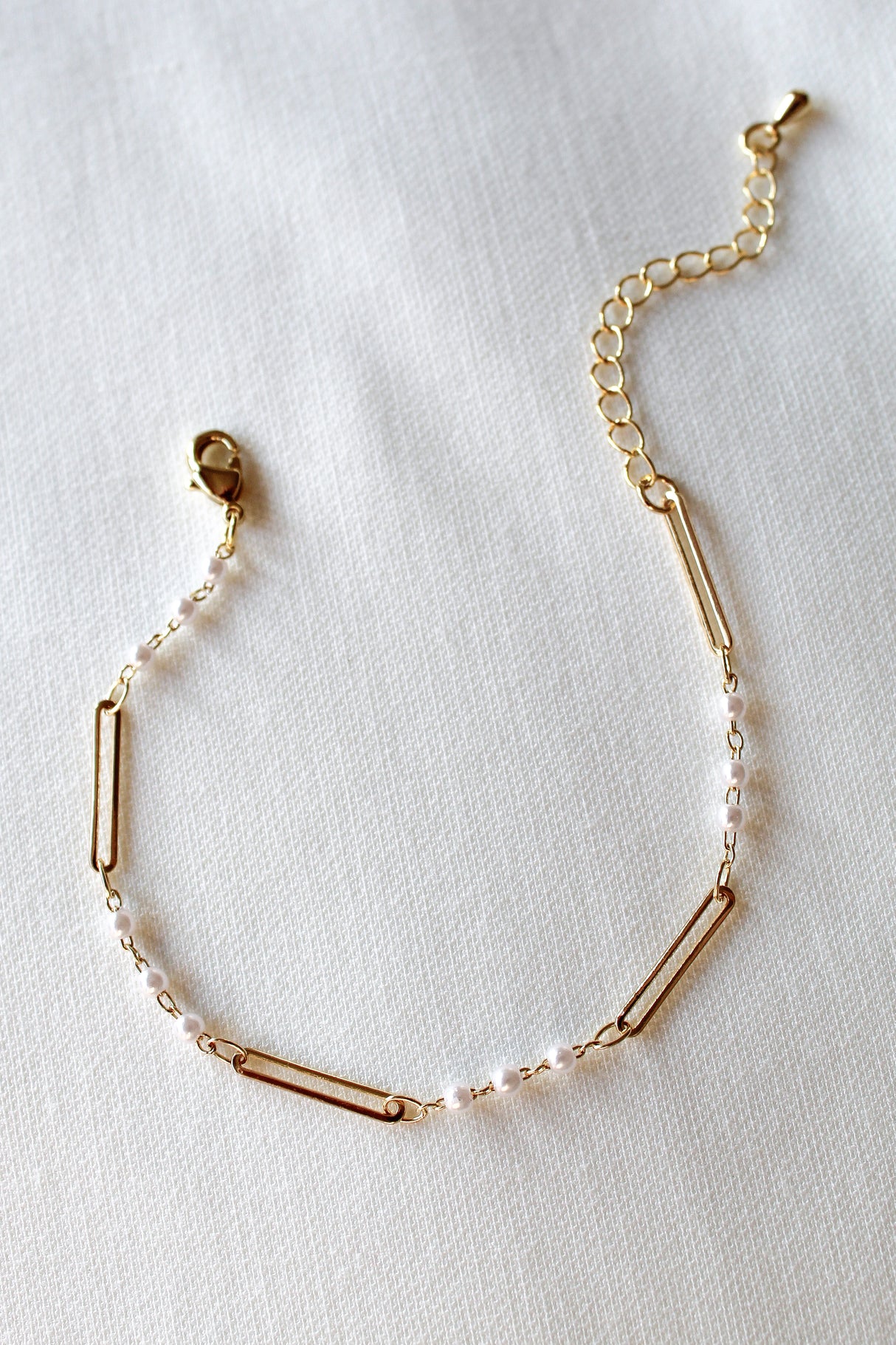 The Marina Pearl Bracelet by Kinsey Designs Archives features pearl-like beads and elongated gold links, showcased on a textured white fabric background.