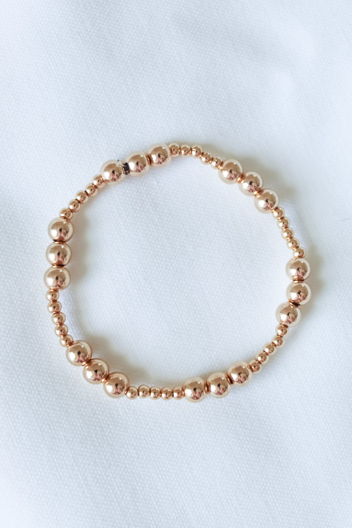 The Mia Bracelet by Kinsey Designs, shown in multiple sizes, features gold-filled beads and an adjustable stretch design, arranged in a circle on a white fabric background.