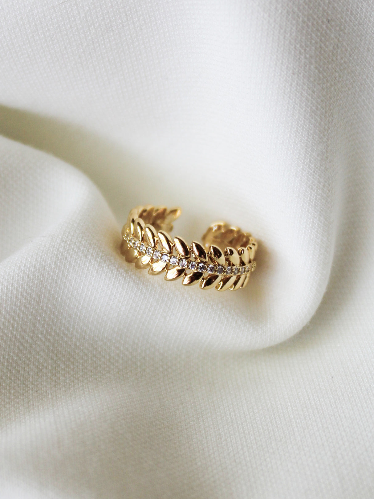 The Nenya Ring by Kinsey Designs features a gold-filled adjustable band adorned with small diamonds in a delicate leaf motif, shown on white textured fabric.