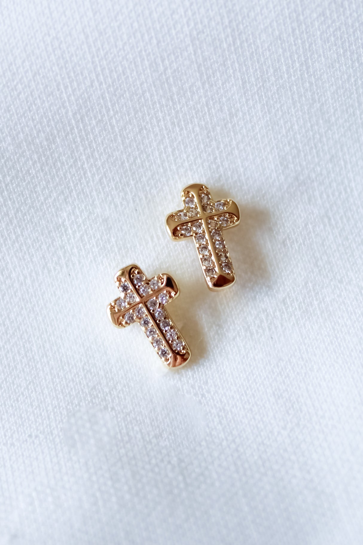 The Noah Cross Studs by Kinsey Designs feature two gold cross earrings adorned with small clear gemstones on a white fabric background, ideal for those who love minimalist or meaningful faith jewelry.
