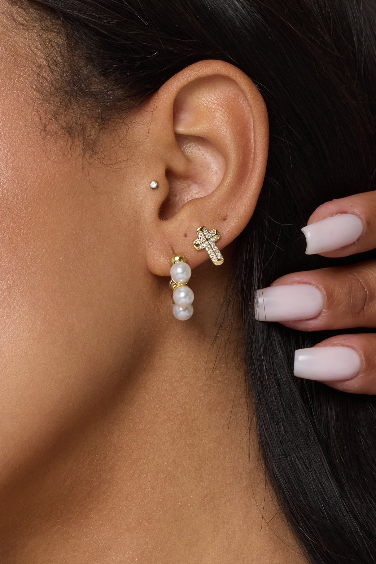 A close-up shows an ear adorned with Kinsey Designs' Noah Cross Studs and pearl hoops, plus a manicured hand nearby.