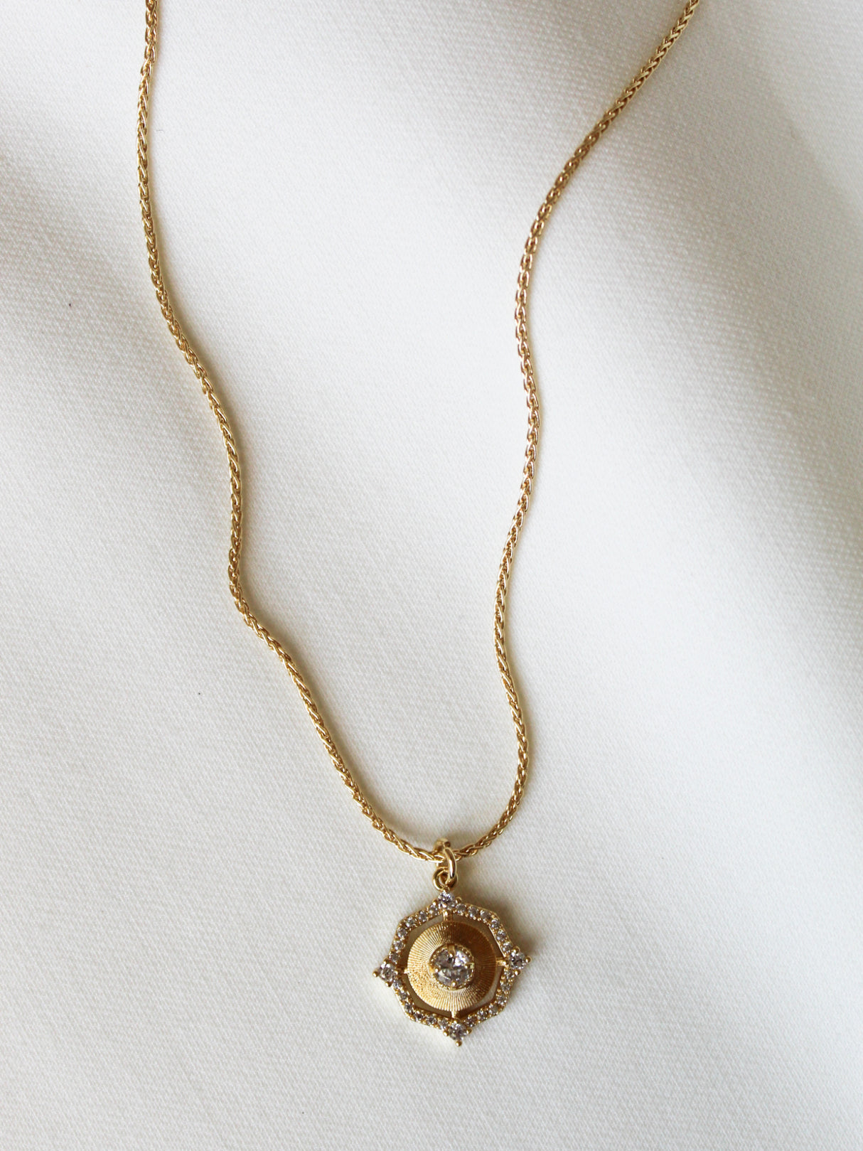 The Orion Detachable Necklace by Kinsey Designs features a round, ornate pendant with a central gemstone, shown on a white fabric background.