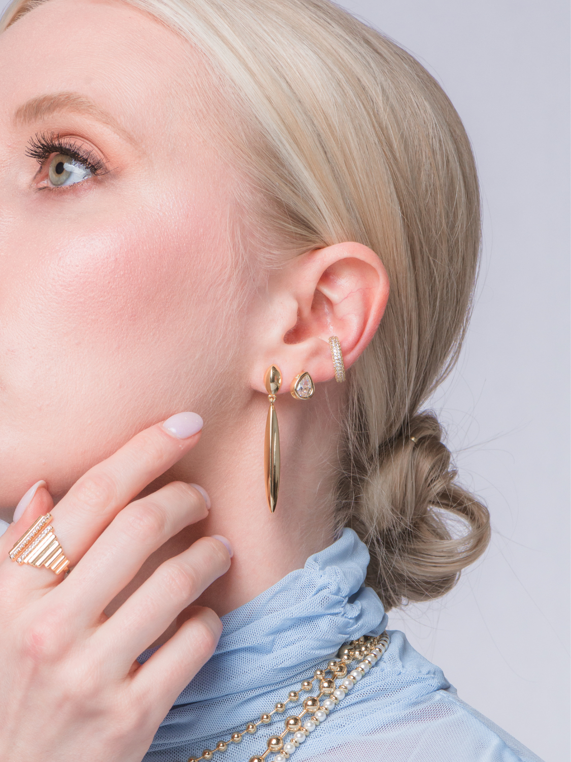 A woman with blonde hair in a low bun wears Kinsey Designs' Pleat Drop Earrings, a gold ring, and a beaded blue top—perfect finds for a sample sale.