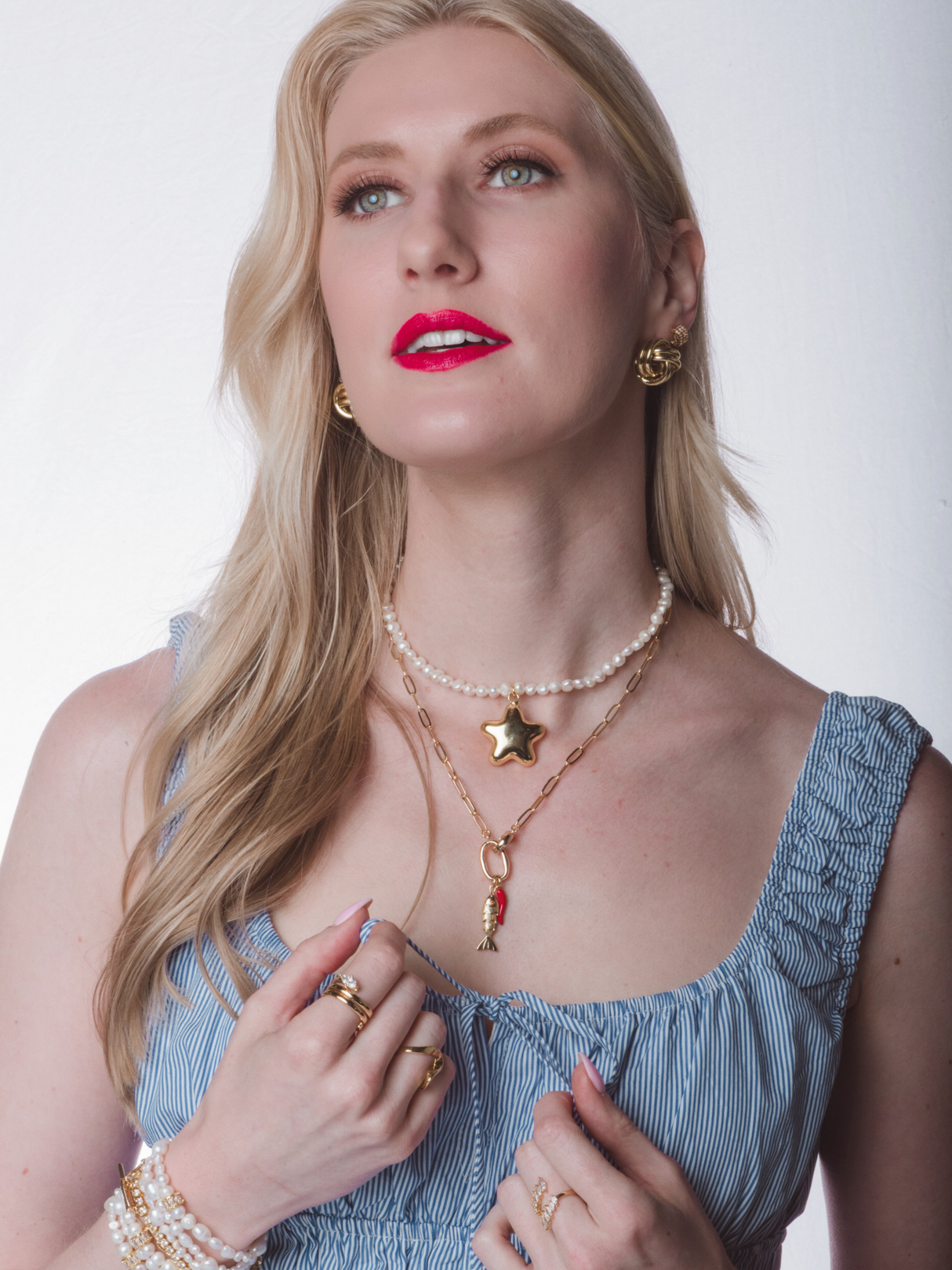 A woman with long blonde hair and red lipstick, wearing a blue dress and the Kinsey Designs Paulette Puffy Star Necklace—a layered gold filled piece with a star pendant—looks upward.