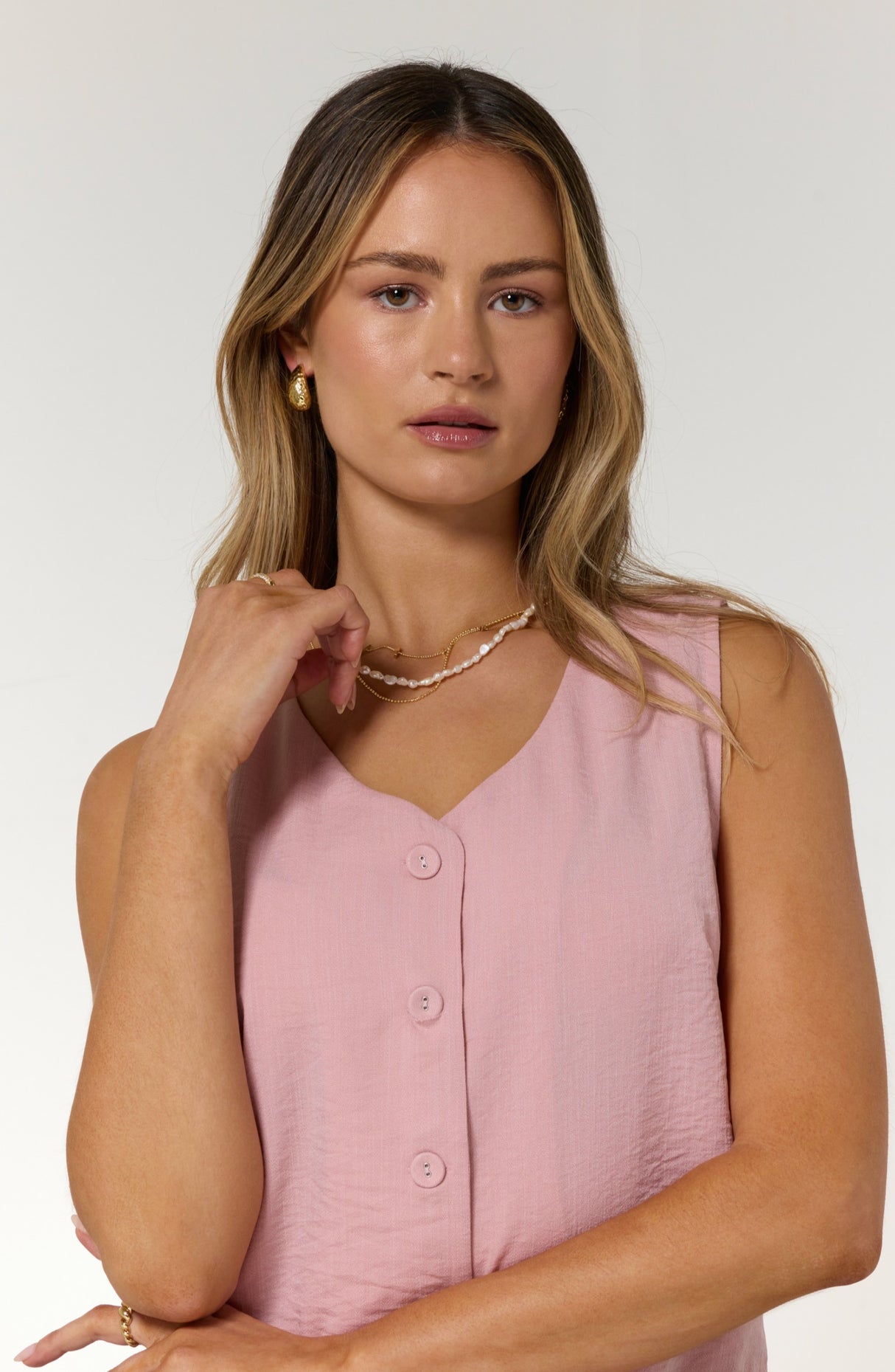 A woman with wavy blonde hair wears a pink sleeveless button-up top and the Kinsey Designs Pearl Layering Necklace, posing against a white background.