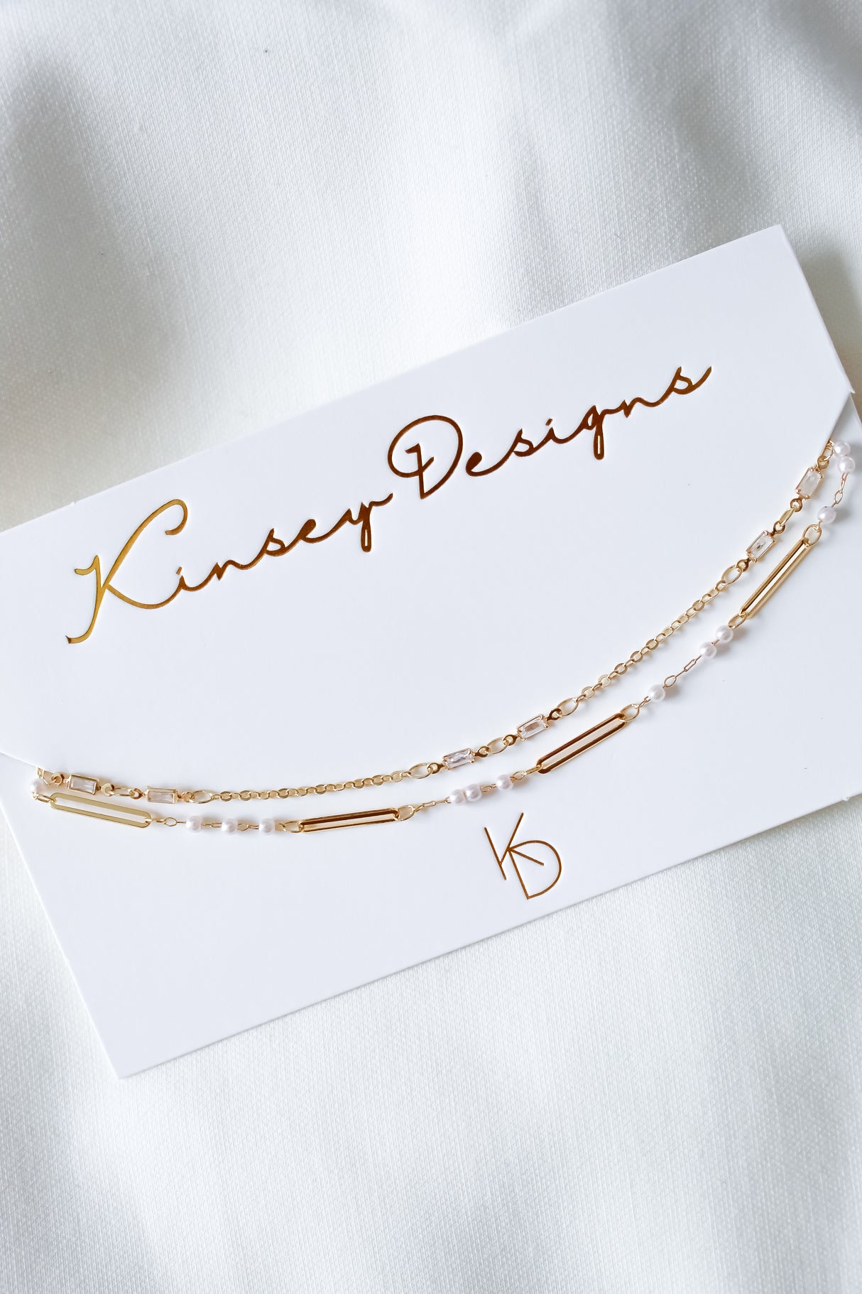 The gold and pearl chain bracelet is displayed on a Kinsey Designs card atop a white fabric background—perfect for pairing with the Kinsey Designs Pelly Layer Necklace from our exclusive sample sale.