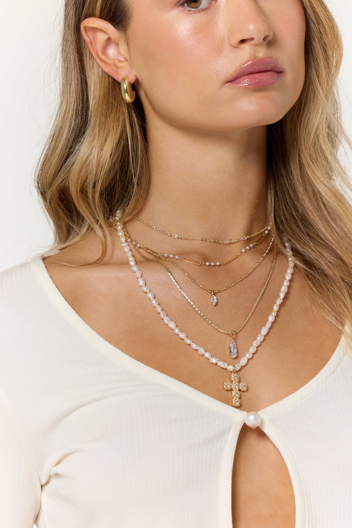 A woman wears the Kinsey Designs Pelly Layer Necklace—gold and pearl strands with a cross pendant—plus gold hoop earrings and a white top, making it the perfect accessory for your next sample sale find.