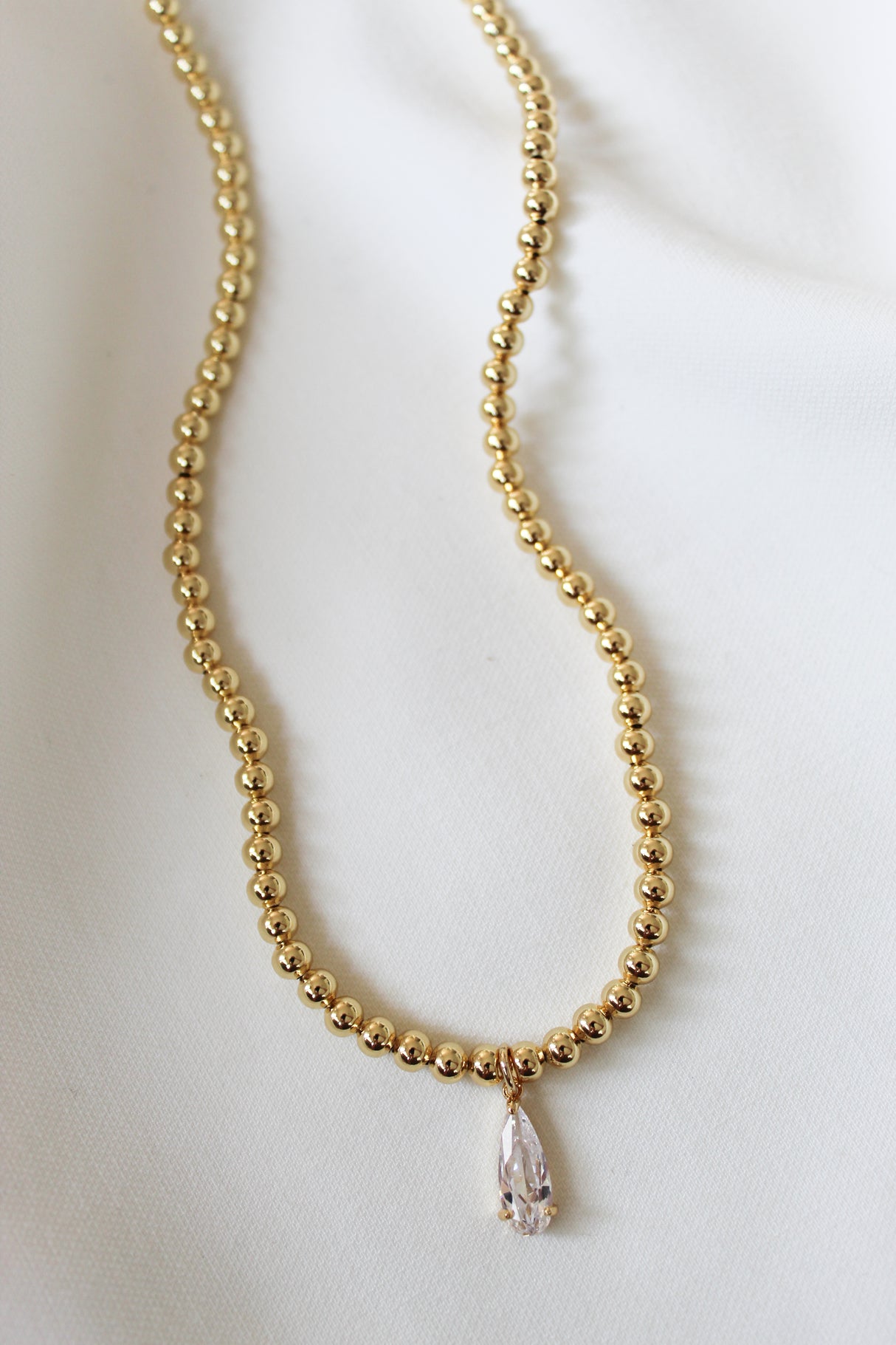The Penn Necklace by Kinsey Designs Archives, featuring gold beads and a pear-shaped pendant, is displayed elegantly on a white fabric background.