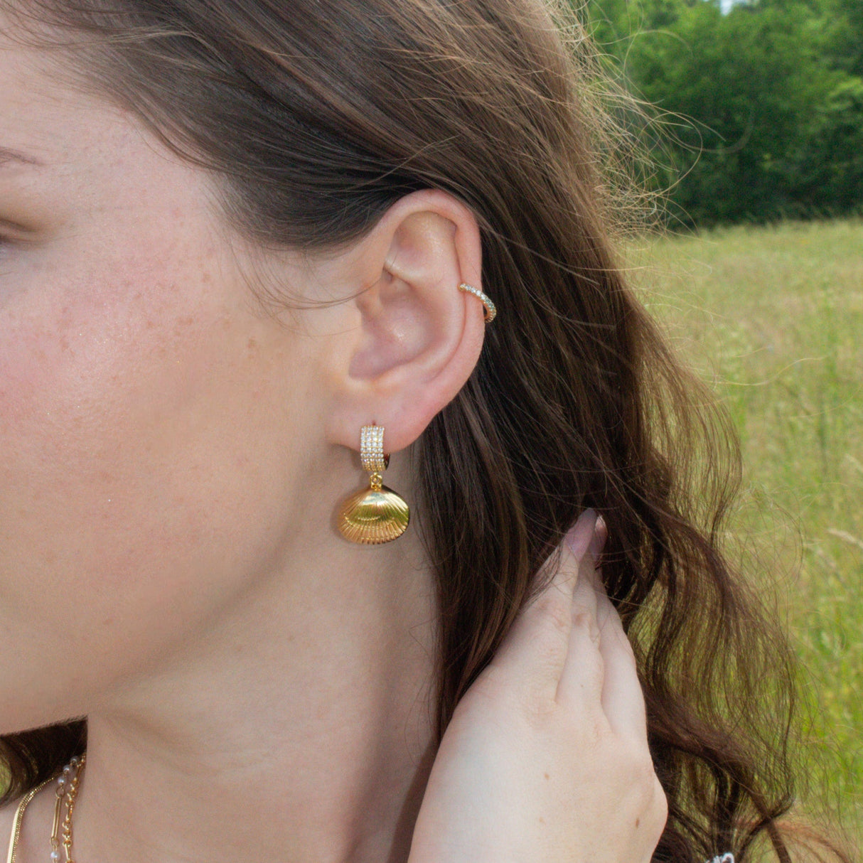 Woman with wavy brown hair wears Kinsey Designs Archives’ Alana Huggies gold filled shell earrings and an ear cuff, hand near ear in a grassy field—ideal for those seeking stunning earrings at our sample sale.