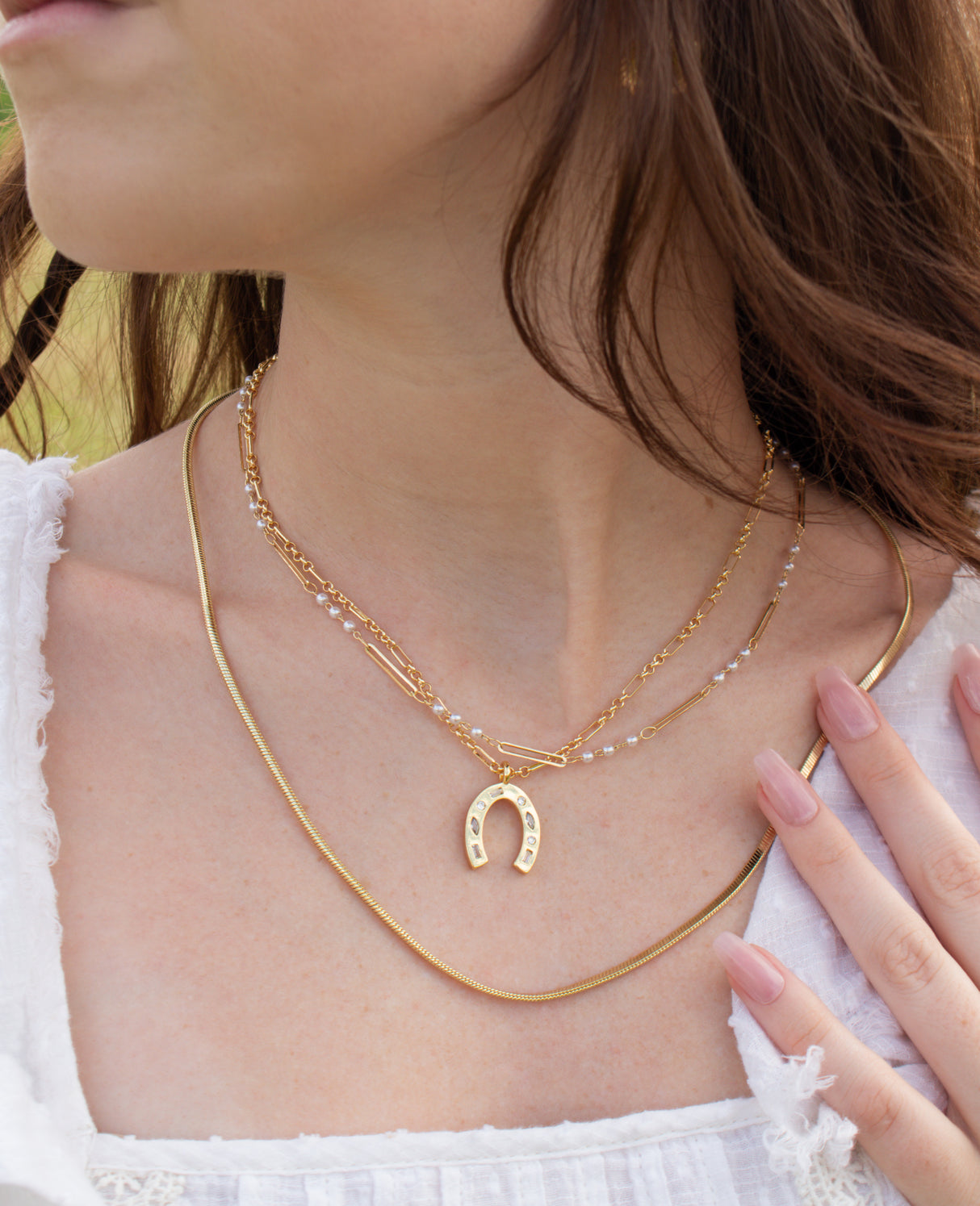 A woman wearing the Kinsey Designs Archives Isla Necklace, a gold filled layered piece with a horseshoe pendant on an 18" chain, paired with a white top and pink manicured nails.