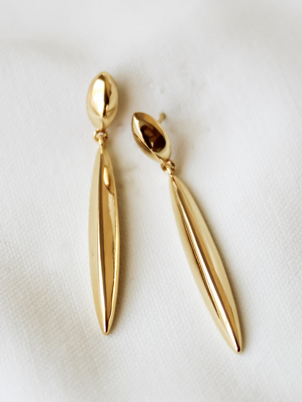 The Kinsey Designs Pleat Drop Earrings feature elongated, leaf-like gold-filled shapes, beautifully displayed on a white fabric background—perfect for discovering at your next sample sale.
