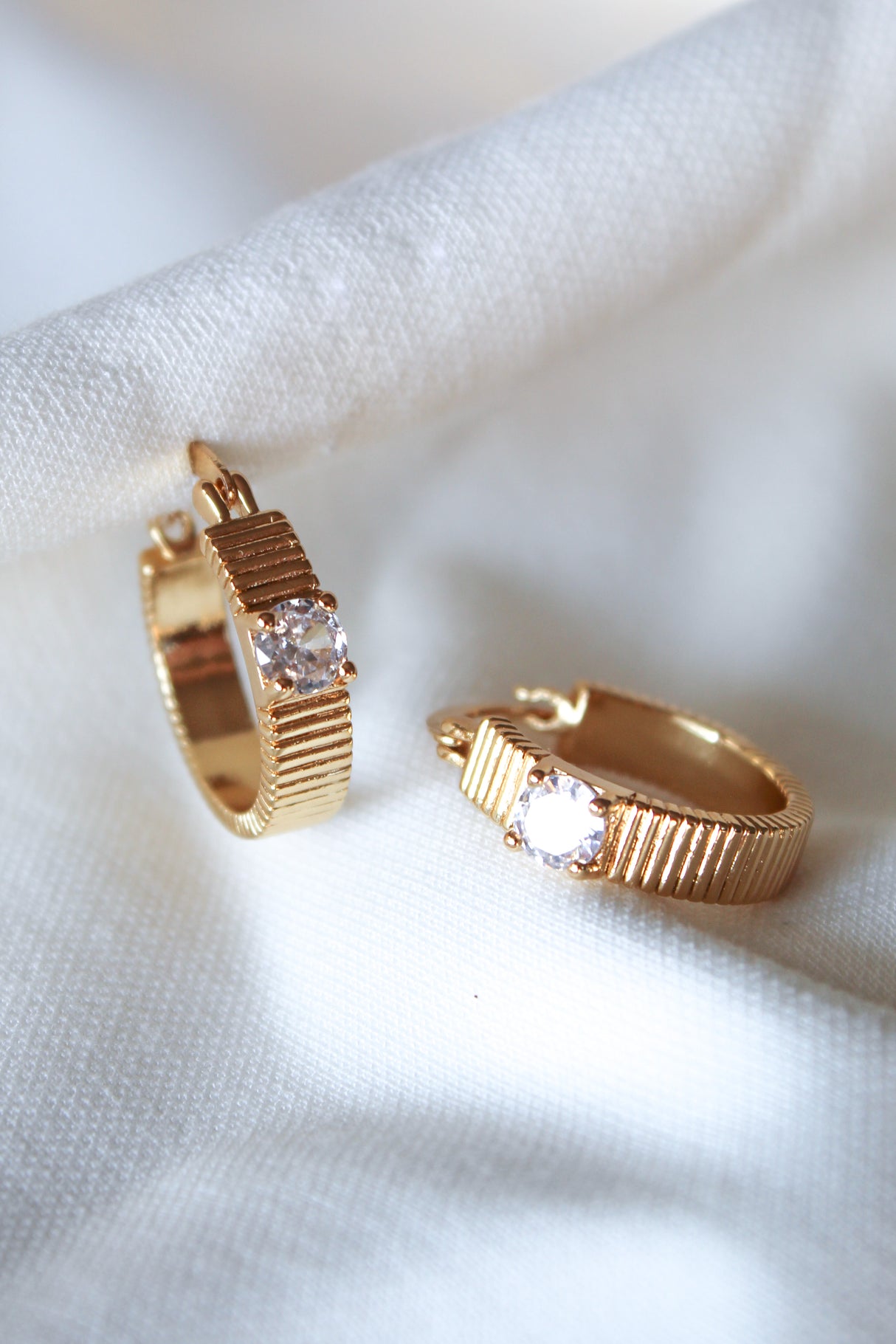 The Quinn Hoop earrings by Kinsey Designs feature gold-filled textured bands and clear gemstones, each with a .75" diameter, shown on white fabric.